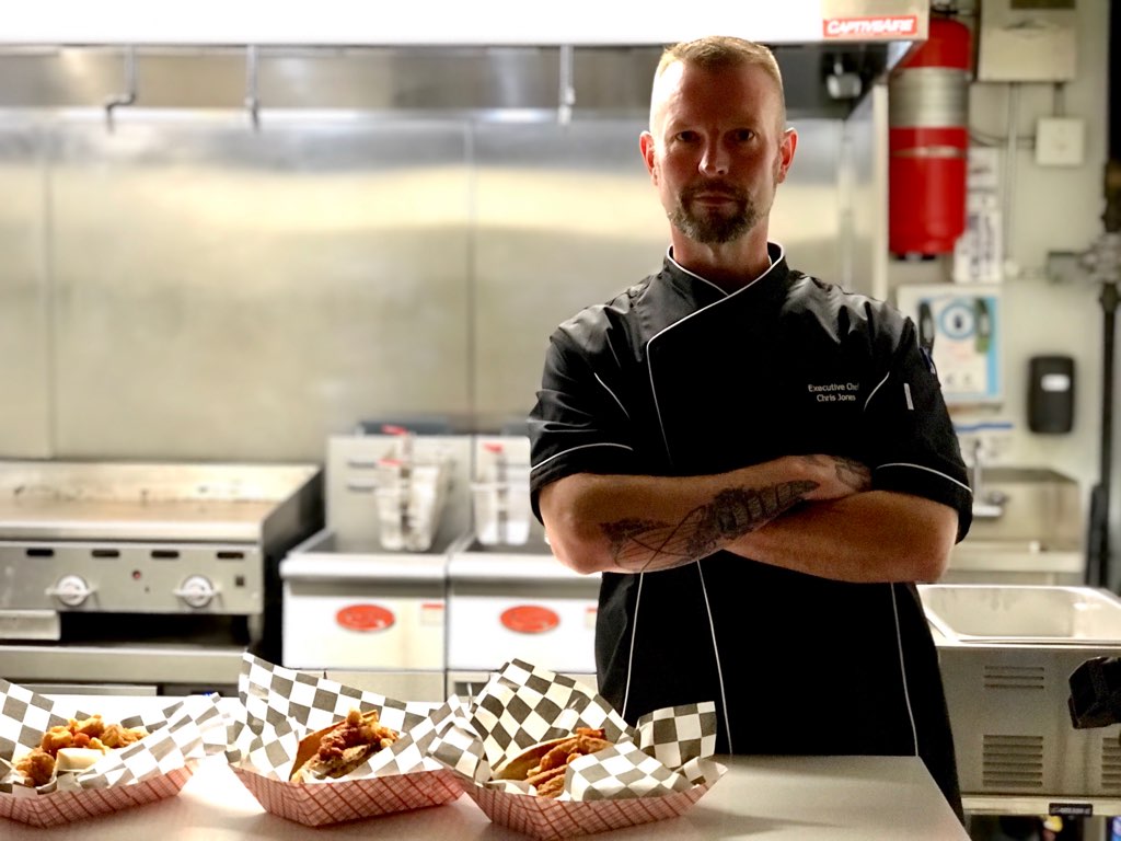 Chef standing with arms crossed in a kitchen, wearing a black uniform, with cooking equipment in the background.