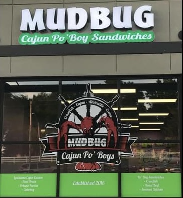 Storefront sign with the name 'MUDBUG Cajun Po' Boy Sandwiches' and a logo featuring a red crawfish, green background, and interior reflections.