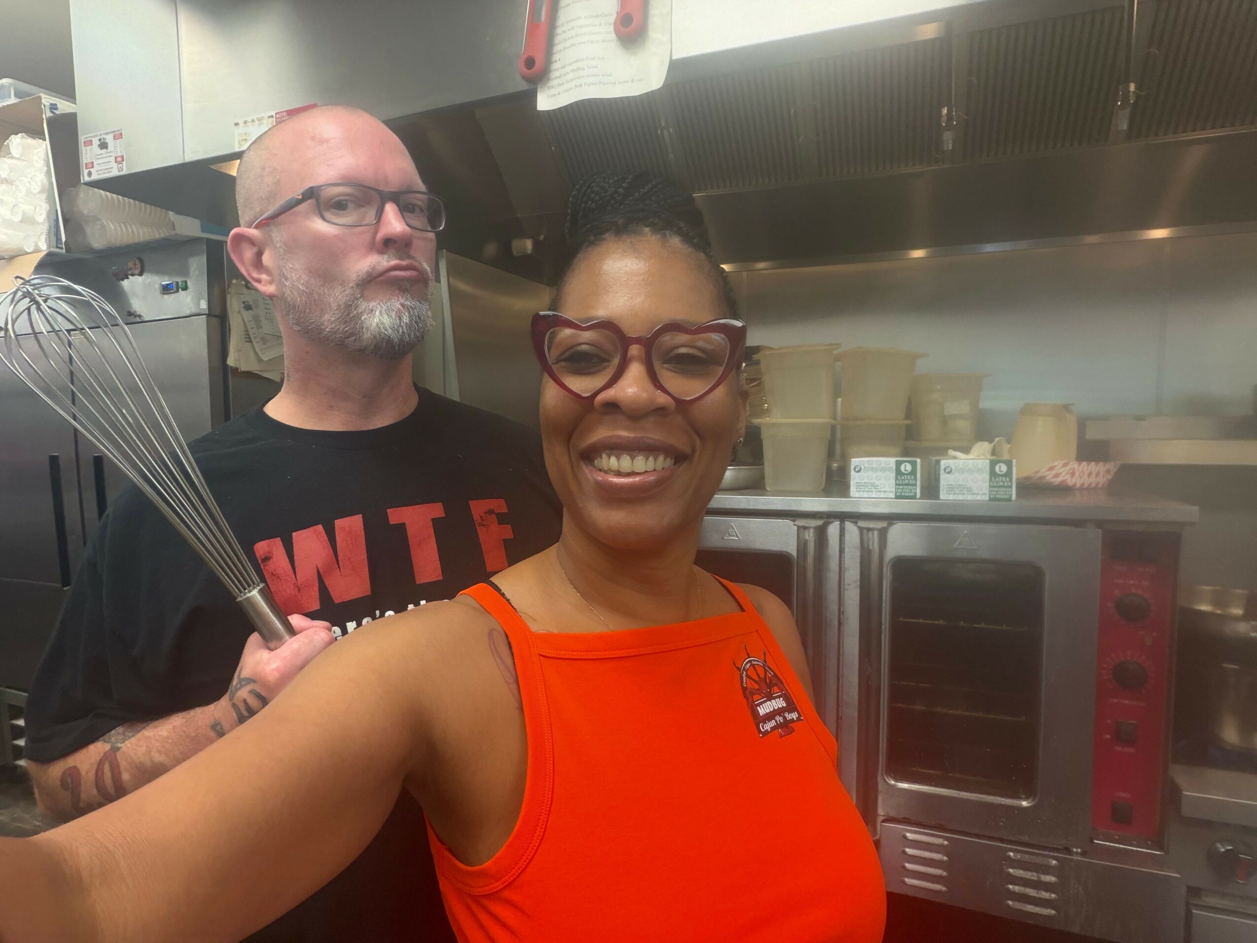 Two people smiling in a kitchen, one wearing glasses and an orange top, the other with a beard and black shirt.
