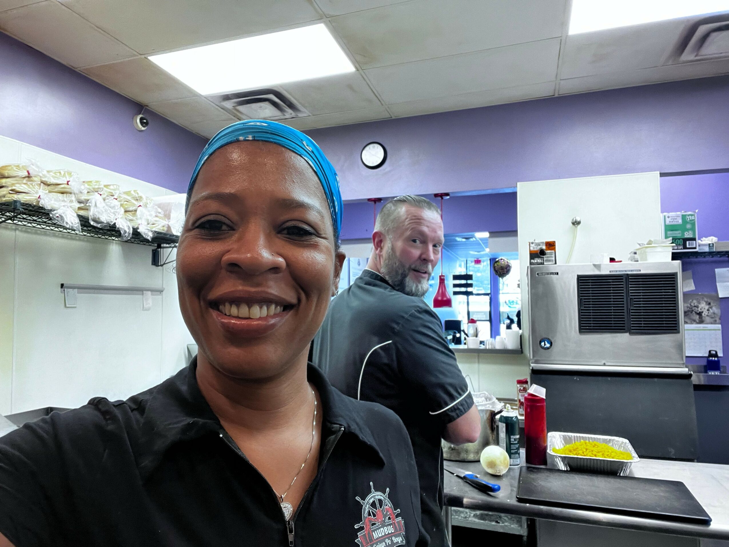 Two people smiling in a laboratory or workspace, one wearing a blue head covering, with equipment and supplies around them.