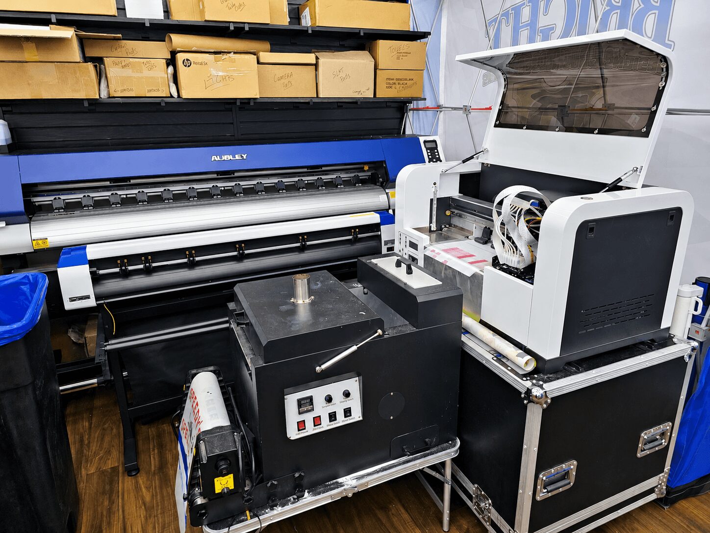 Large digital printing or cutting machine with open cover, control panel, and nearby equipment in a workspace.
