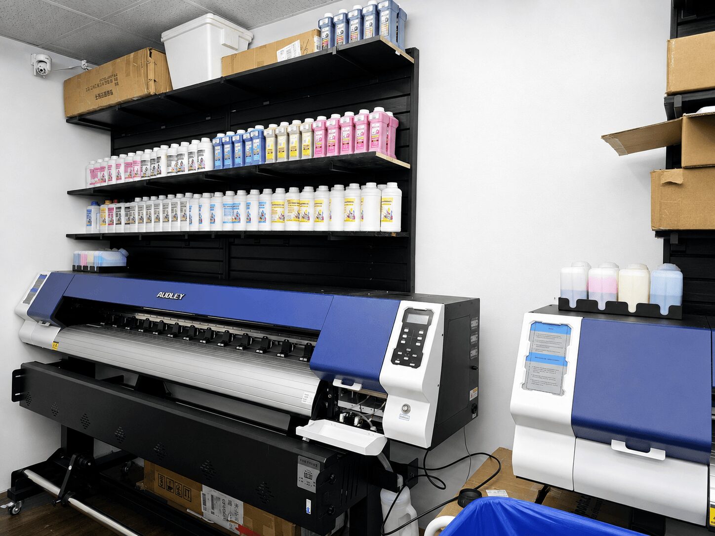 Printer with shelves of colorful bottles above, and boxes on the right side, in a storage room.