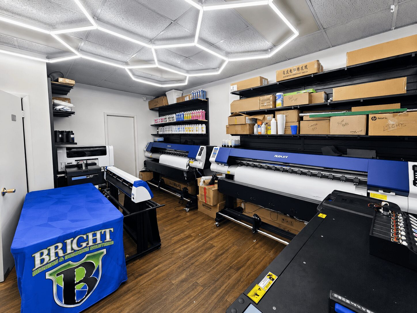Room with large printers, shelves with boxes and supplies, and a table with a blue cover labeled 'BRIGHT'.