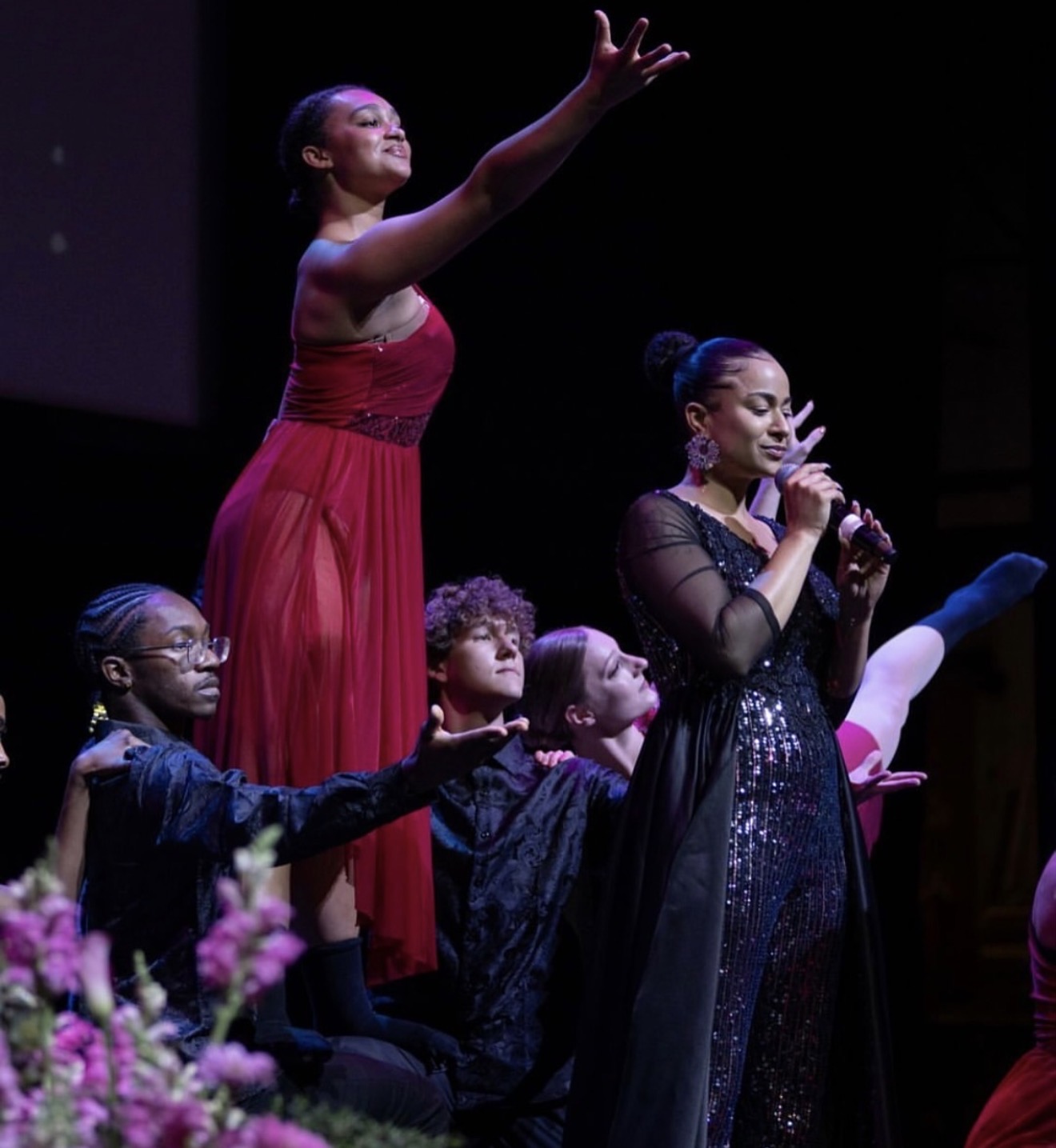 Group of performers on stage, one singing into a microphone, others with arms raised, pink flowers in foreground.