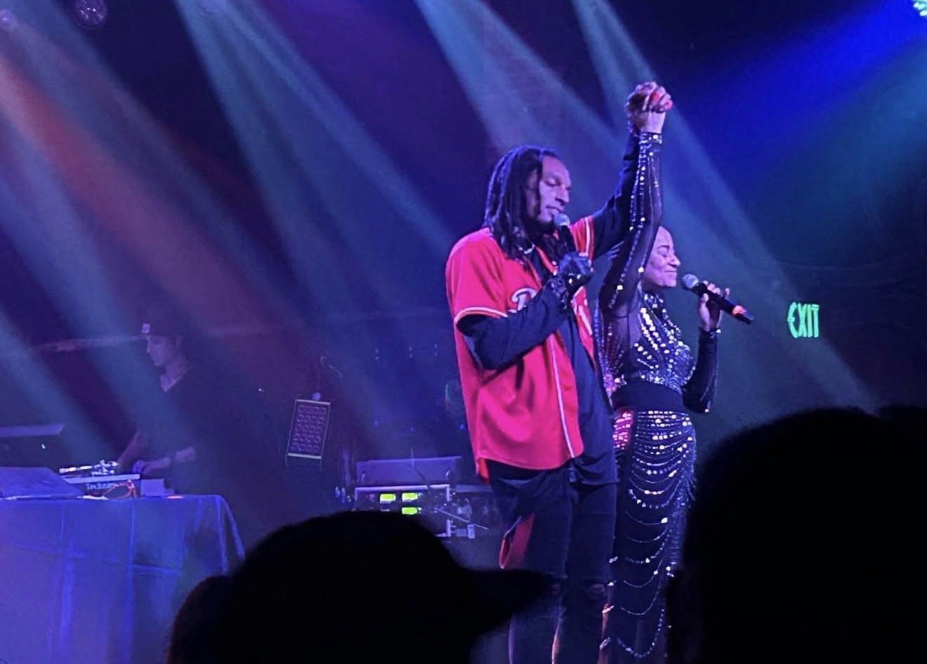 Two performers on stage, one with a microphone and the other raising a hand, under colorful stage lights.