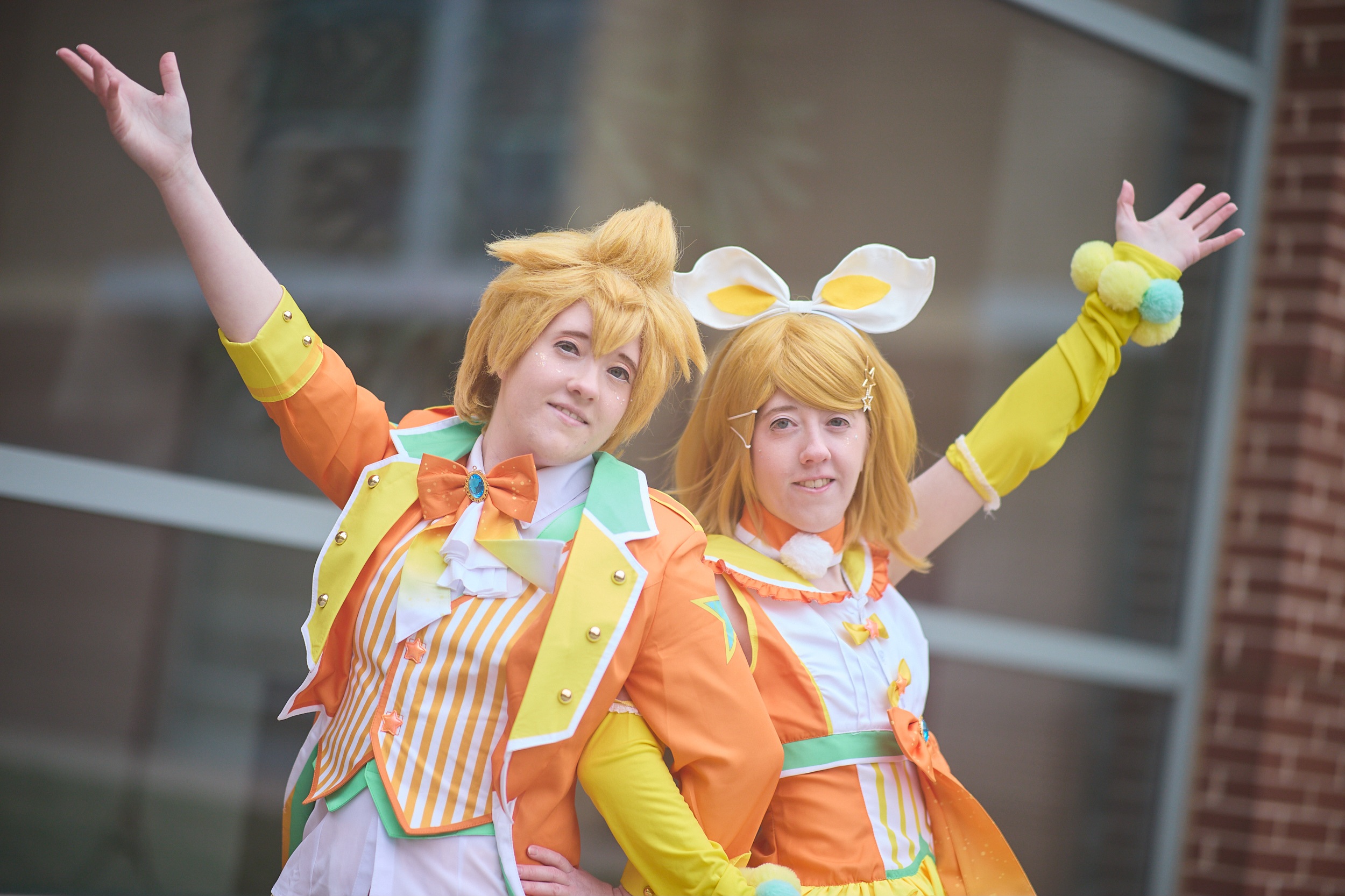 Two people in colorful costumes pose outdoors with arms raised, smiling, in front of a building with glass windows.