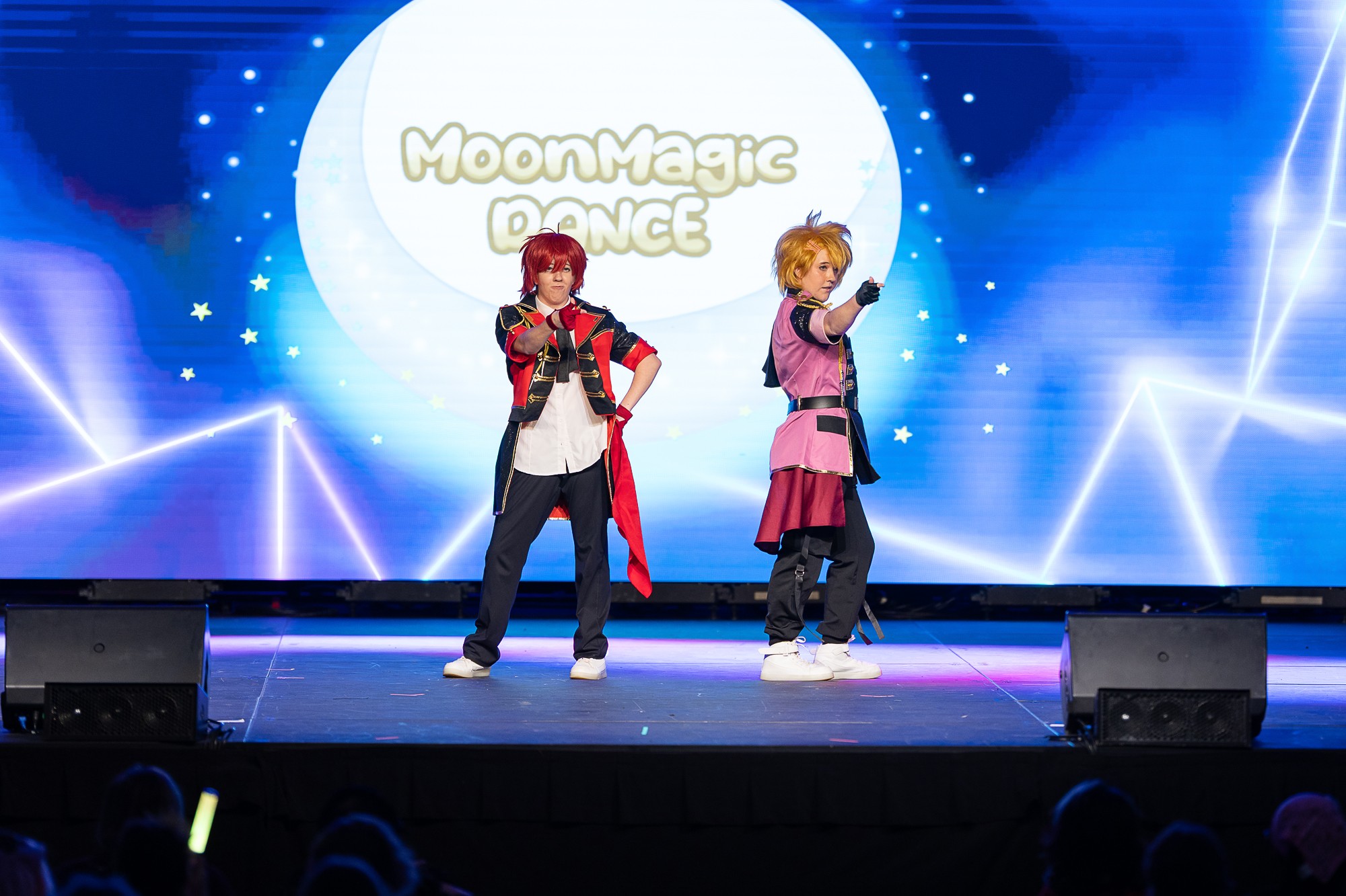 Two performers on stage with a large moon and starry background, one singing into a microphone, the other gesturing.