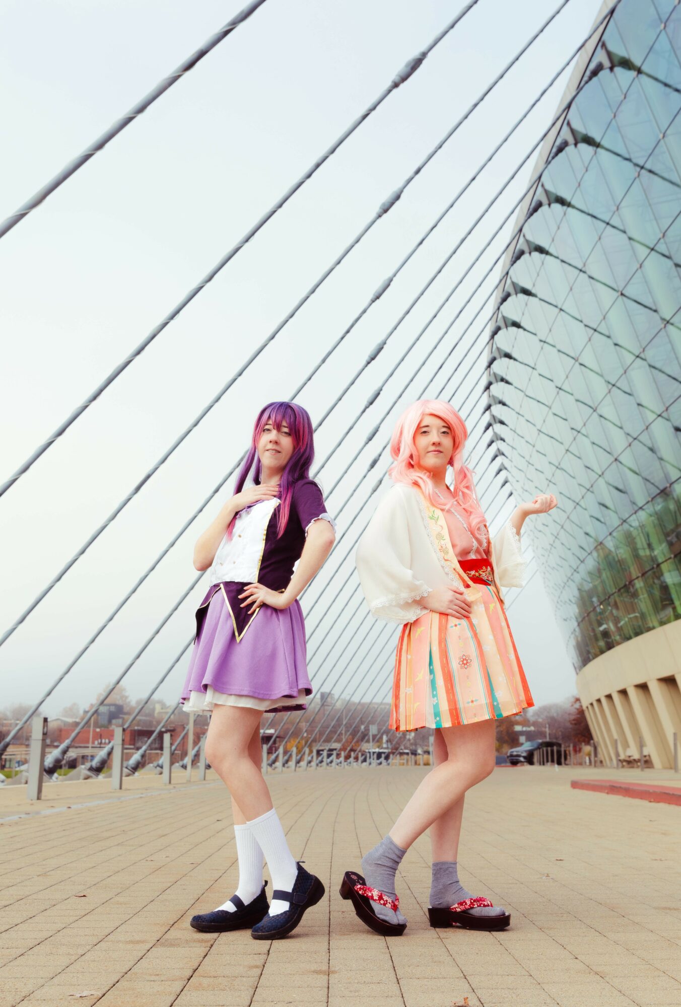 Two young women with colorful hair and fashionable outfits stand outdoors in front of a modern architectural building with glass panels.