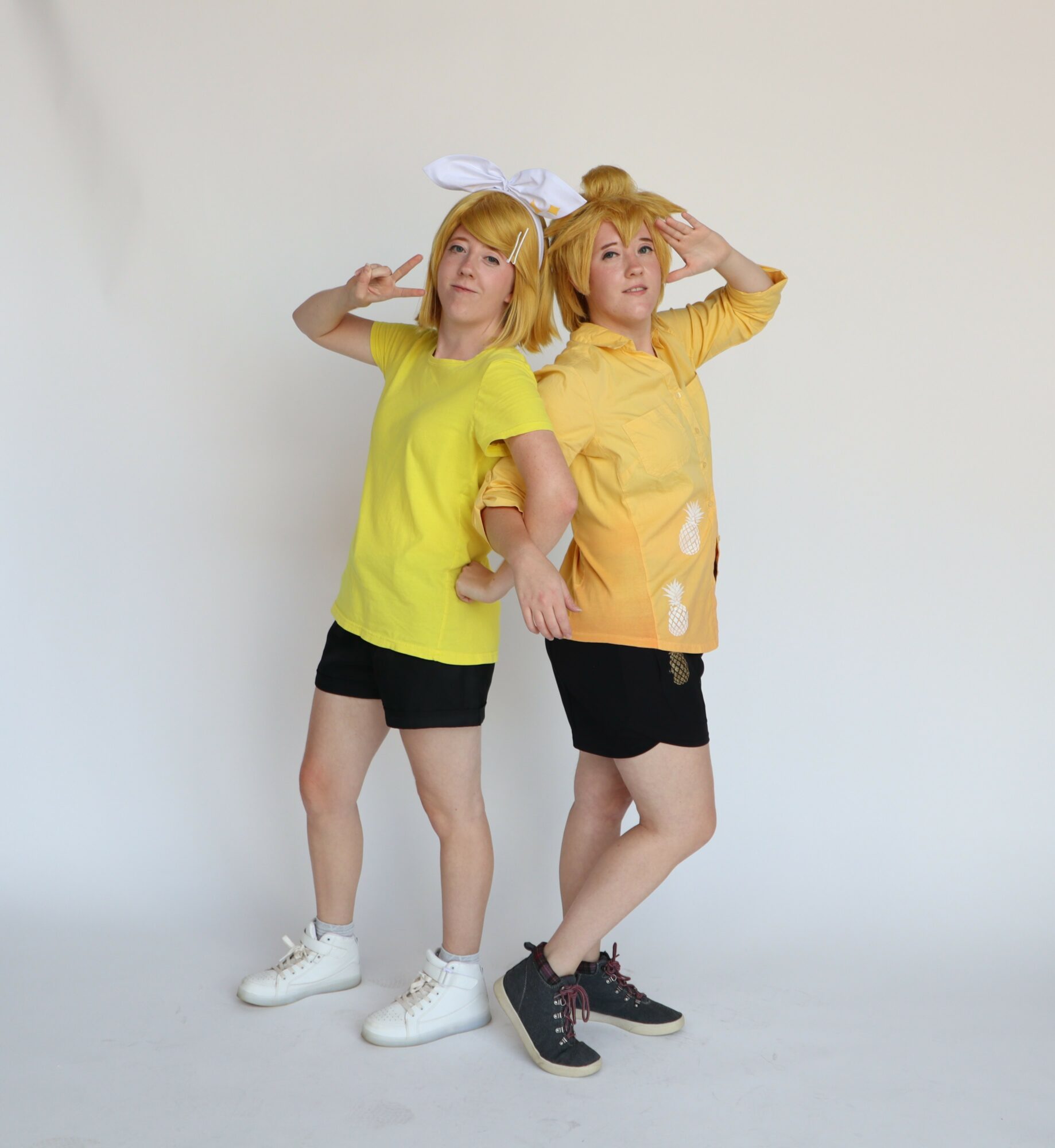 Two young women with blonde hair pose against a plain white background, wearing yellow tops and black shorts, one with a white headband.