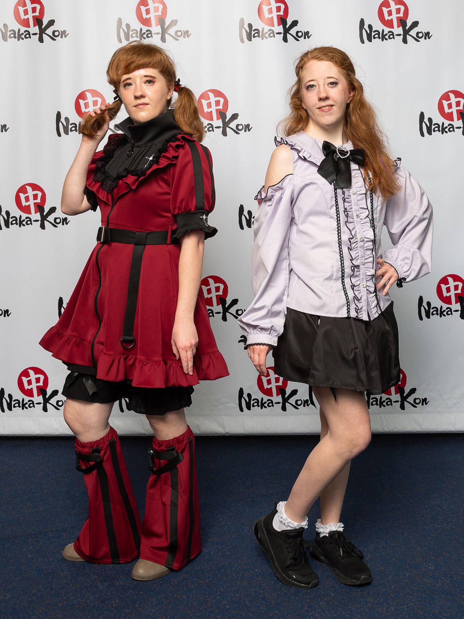 Two young women pose in front of a backdrop with logos, one in a red dress with black accents, the other in a white blouse and black skirt.