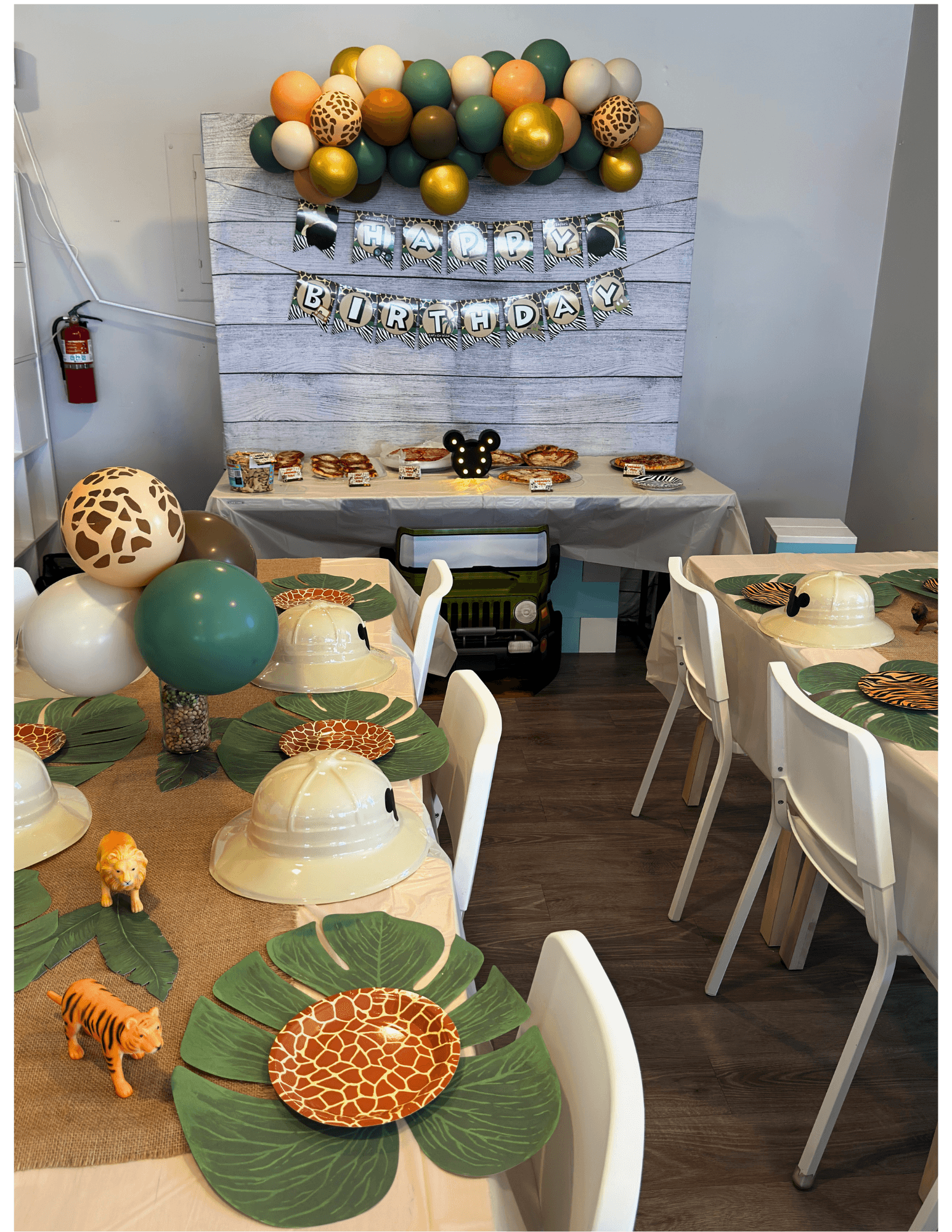 Party setup with tables, chairs, and a decorated backdrop with balloons and a banner, featuring animal-themed table settings.