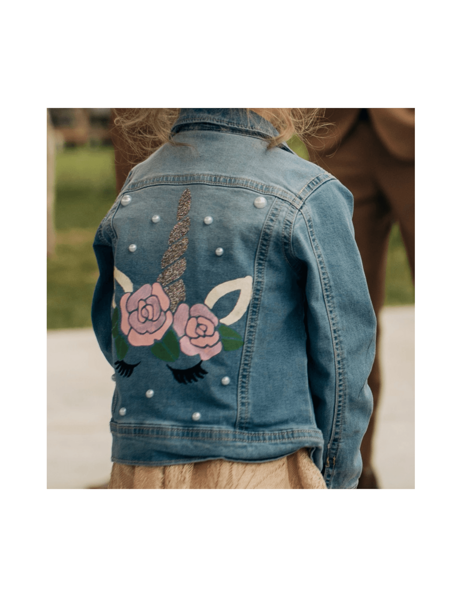 Child wearing a denim jacket with a unicorn and floral design on the back, outdoors. Word 'LOVE' is visible on the jacket.