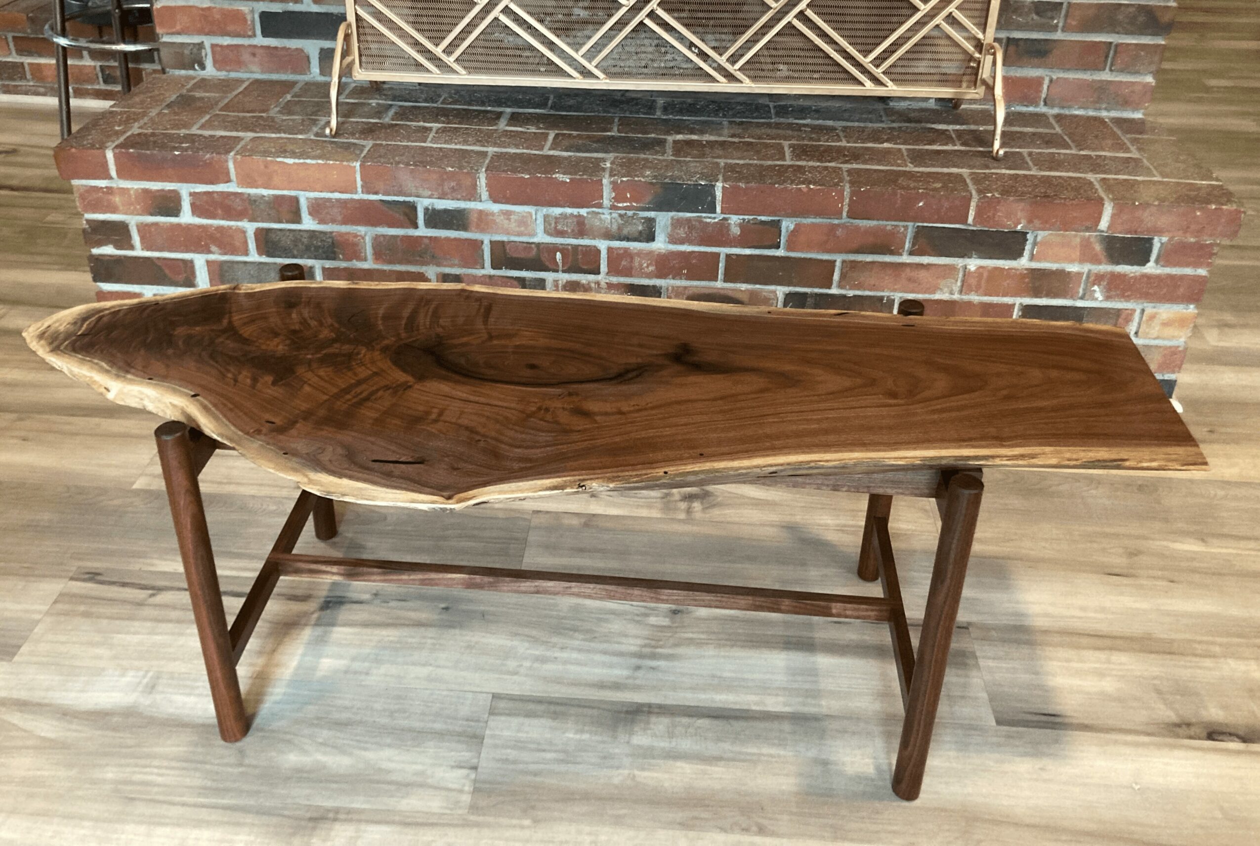 Long wooden table with natural edges, supported by four wooden legs, placed on a wooden floor in front of a brick wall.
