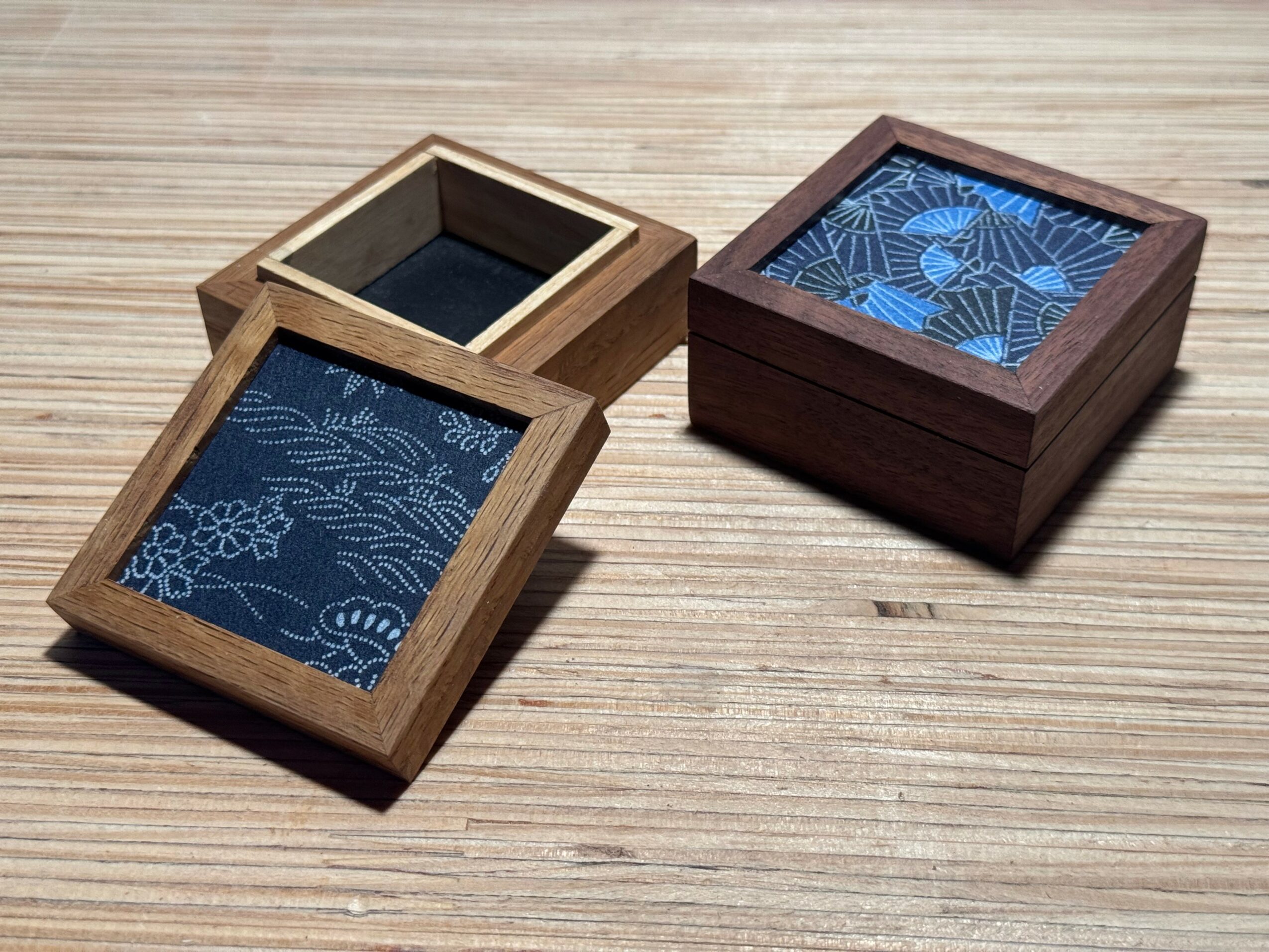 Two wooden boxes with glass tops, one open and one closed, on a wooden surface. The open box shows a blue patterned interior.
