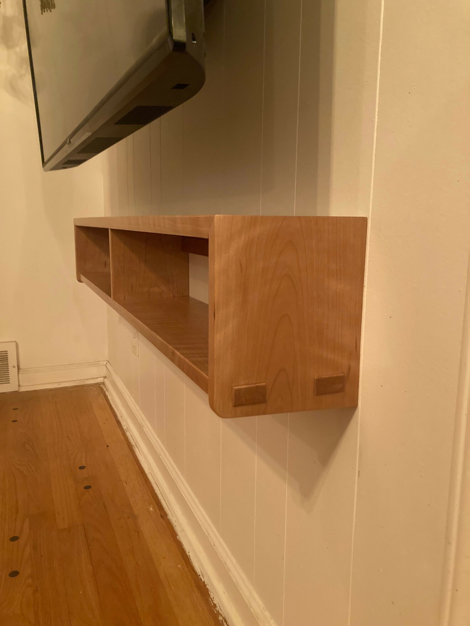 Wooden shelf mounted on a white wall, with a section of a black appliance visible on the left.