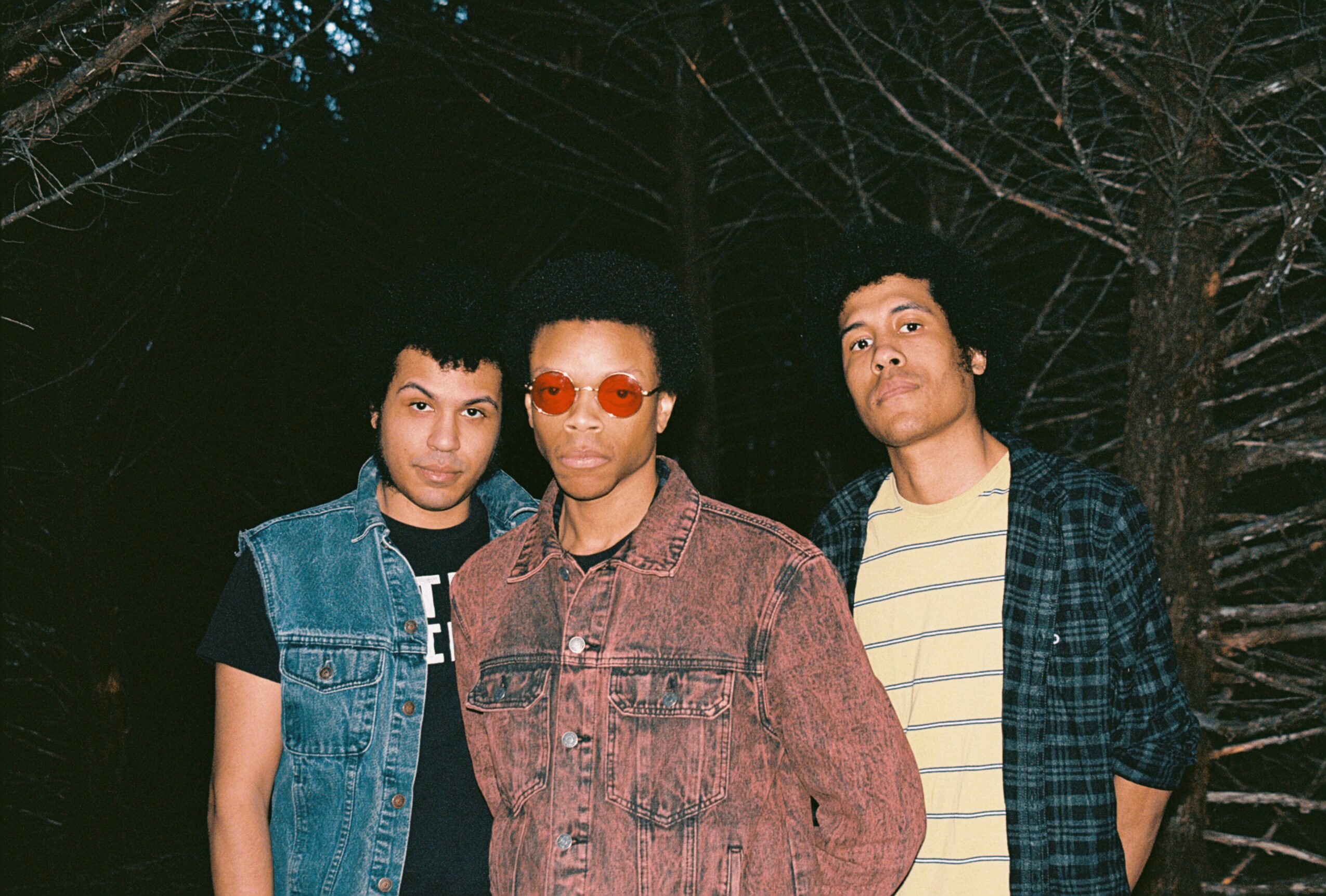 Three young men stand outdoors at night with trees in the background.