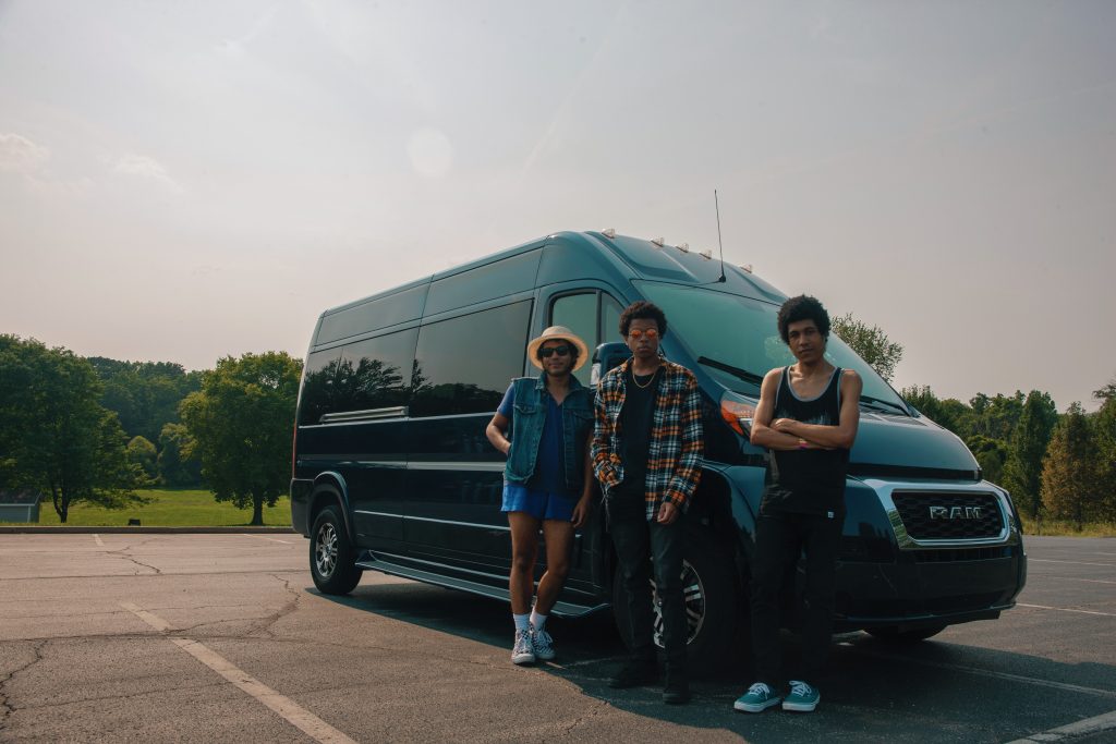 Three people stand in front of a large black van in an open parking lot with trees in the background.