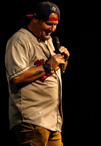 Man wearing a baseball jersey and cap, holding a microphone, smiling on stage against a black background.