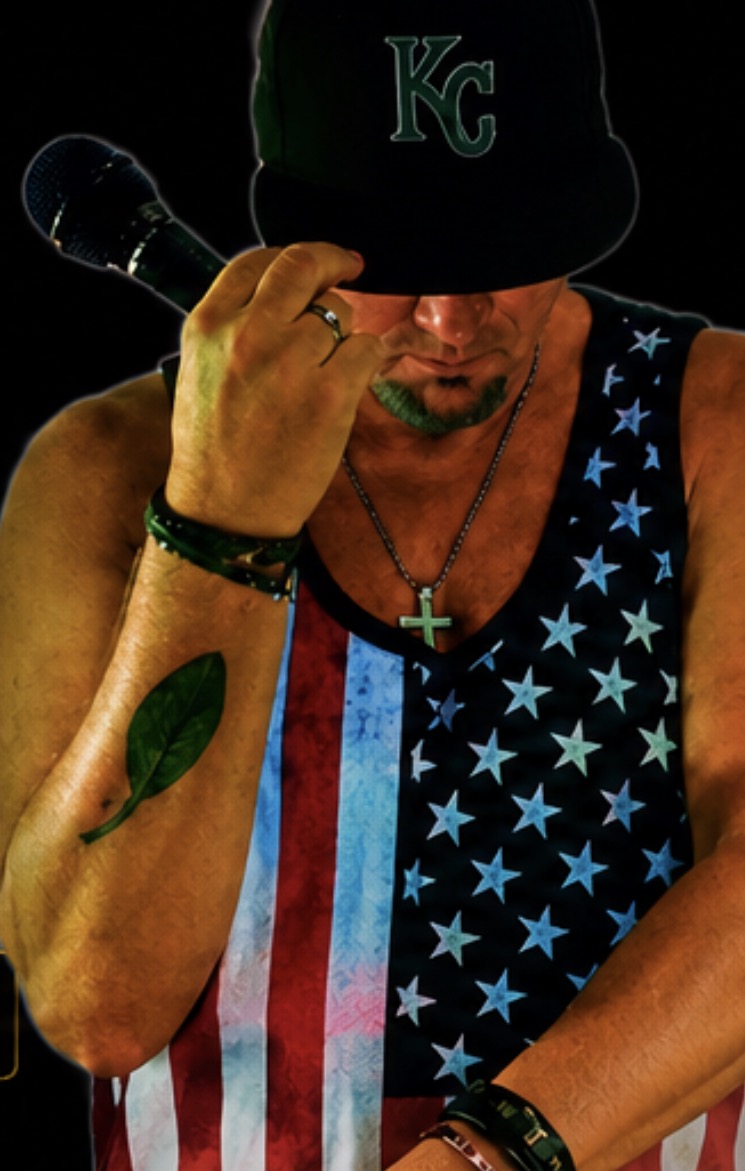 Man wearing a Kansas City cap, sleeveless shirt with American flag design, holding a microphone, with a tattoo on his arm.