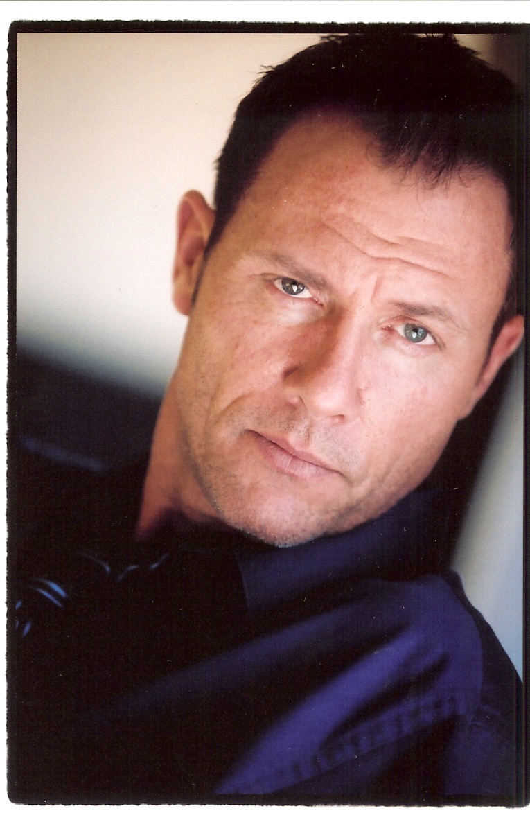 Close-up of a man with short dark hair and blue eyes, looking at the camera with a neutral expression.