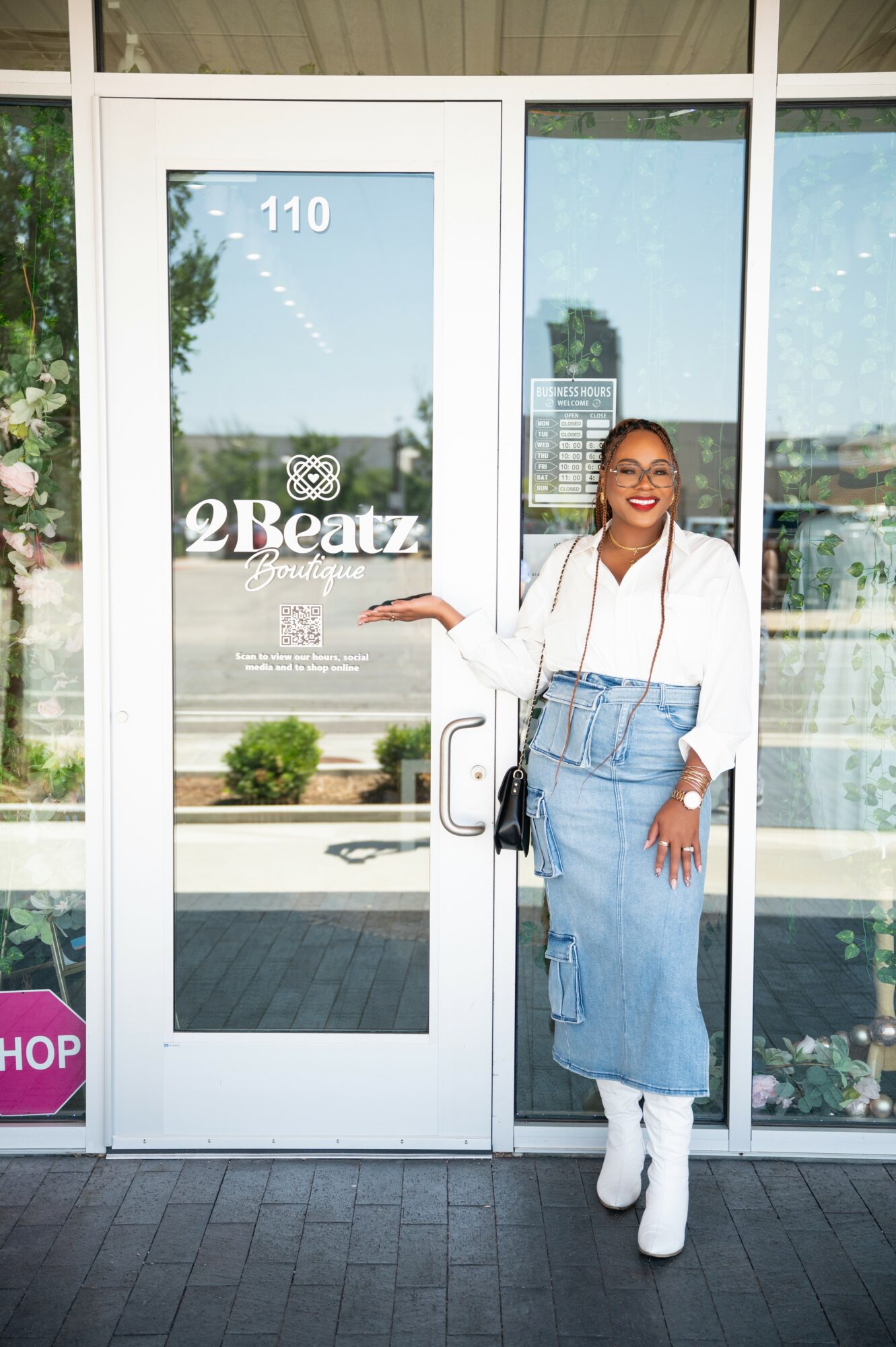 Woman standing outside glass door, smiling, wearing white top, blue jeans, white boots, holding a phone, sunny day.