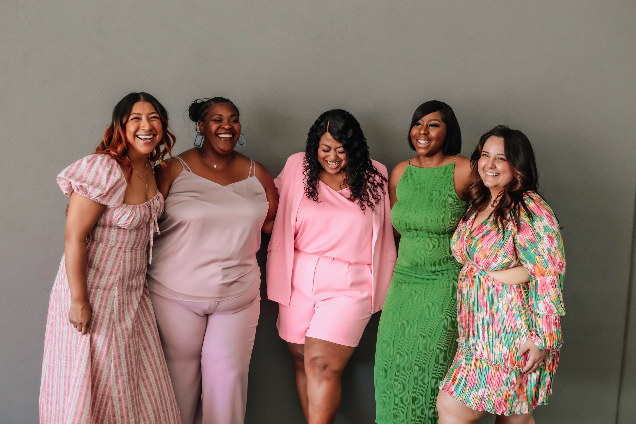 Five women standing together, smiling, wearing colorful dresses and outfits, against a plain background.