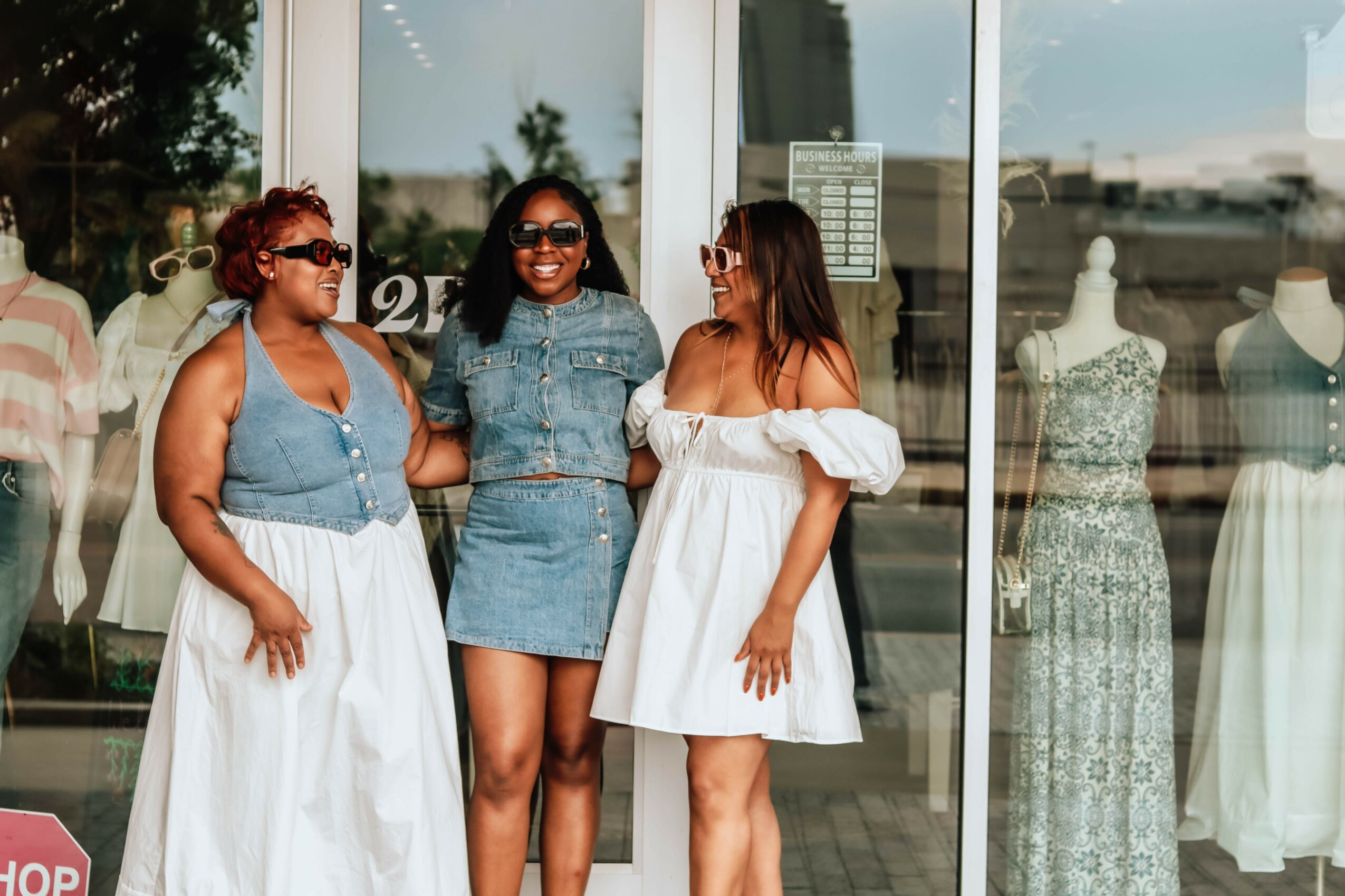 Three women stand outside a store, talking, with mannequins and dresses visible through the glass window behind them.