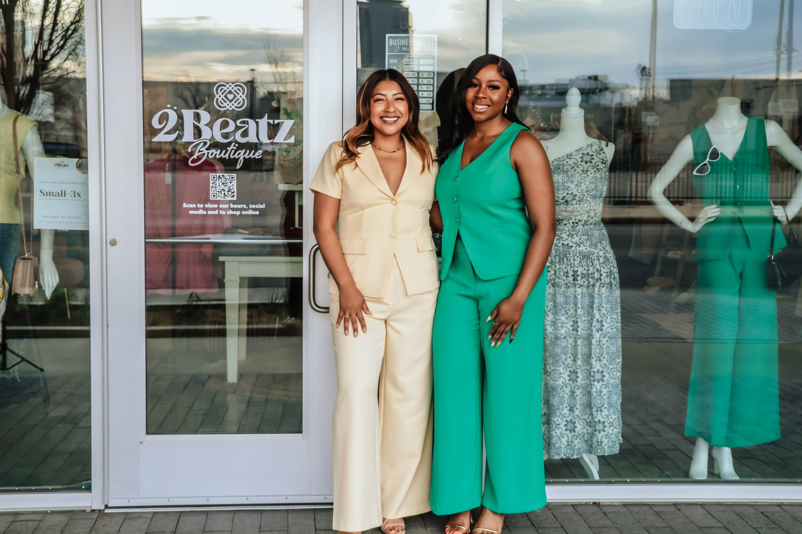 Two women stand outside a boutique store, smiling, with mannequins and dresses visible through the glass window behind them.