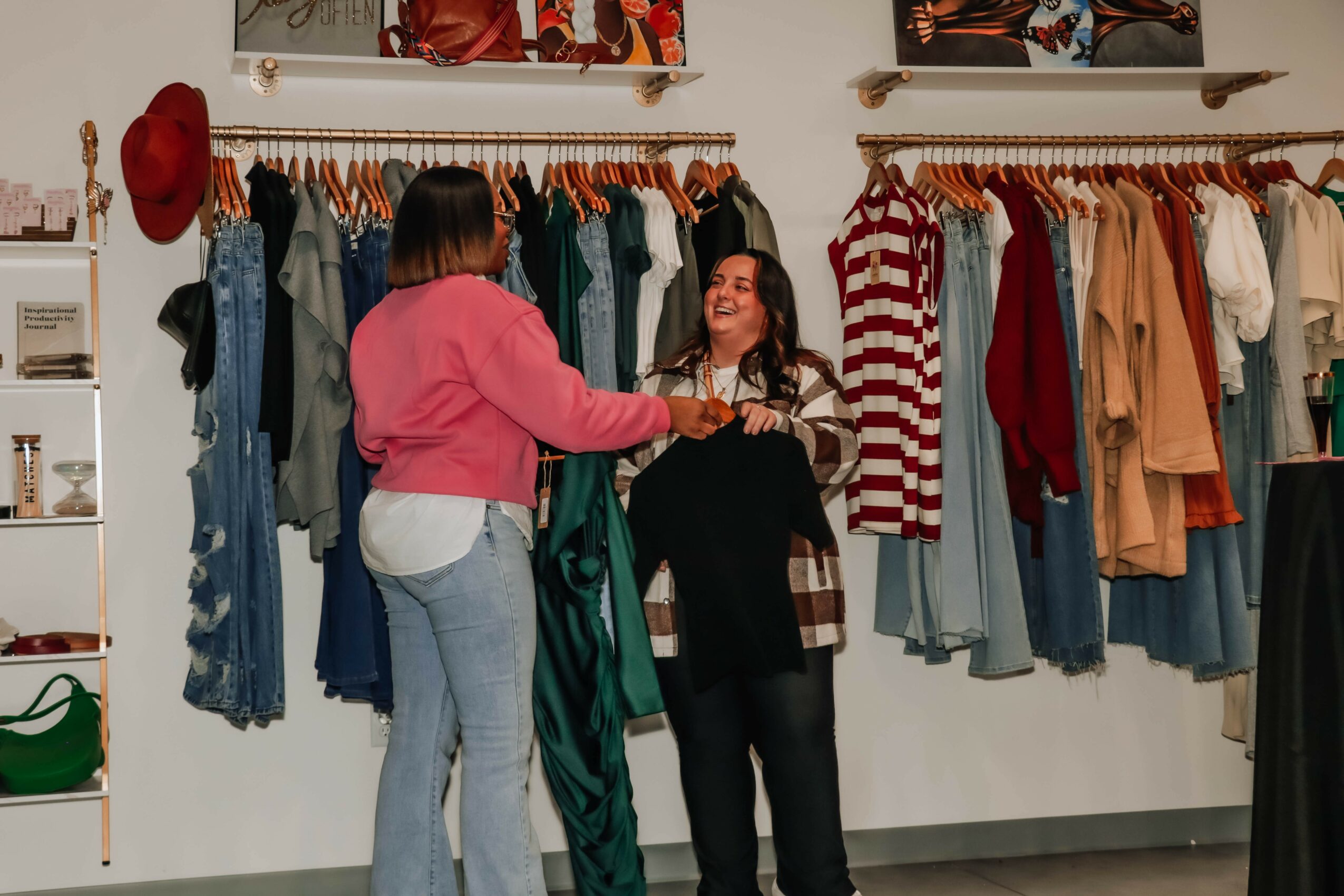 Two women shopping in a clothing store, smiling and holding hands, surrounded by racks of clothes.