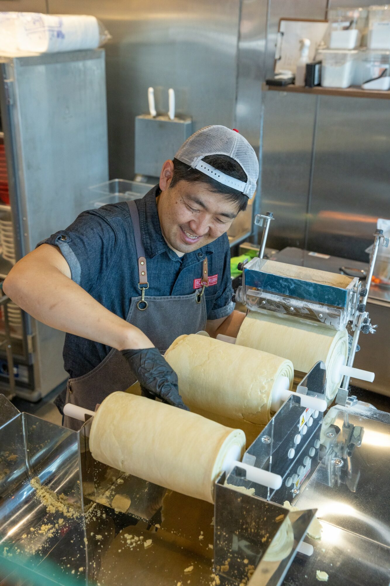 Person wearing a cap and apron rolls dough in a commercial kitchen, smiling while working at a machine.