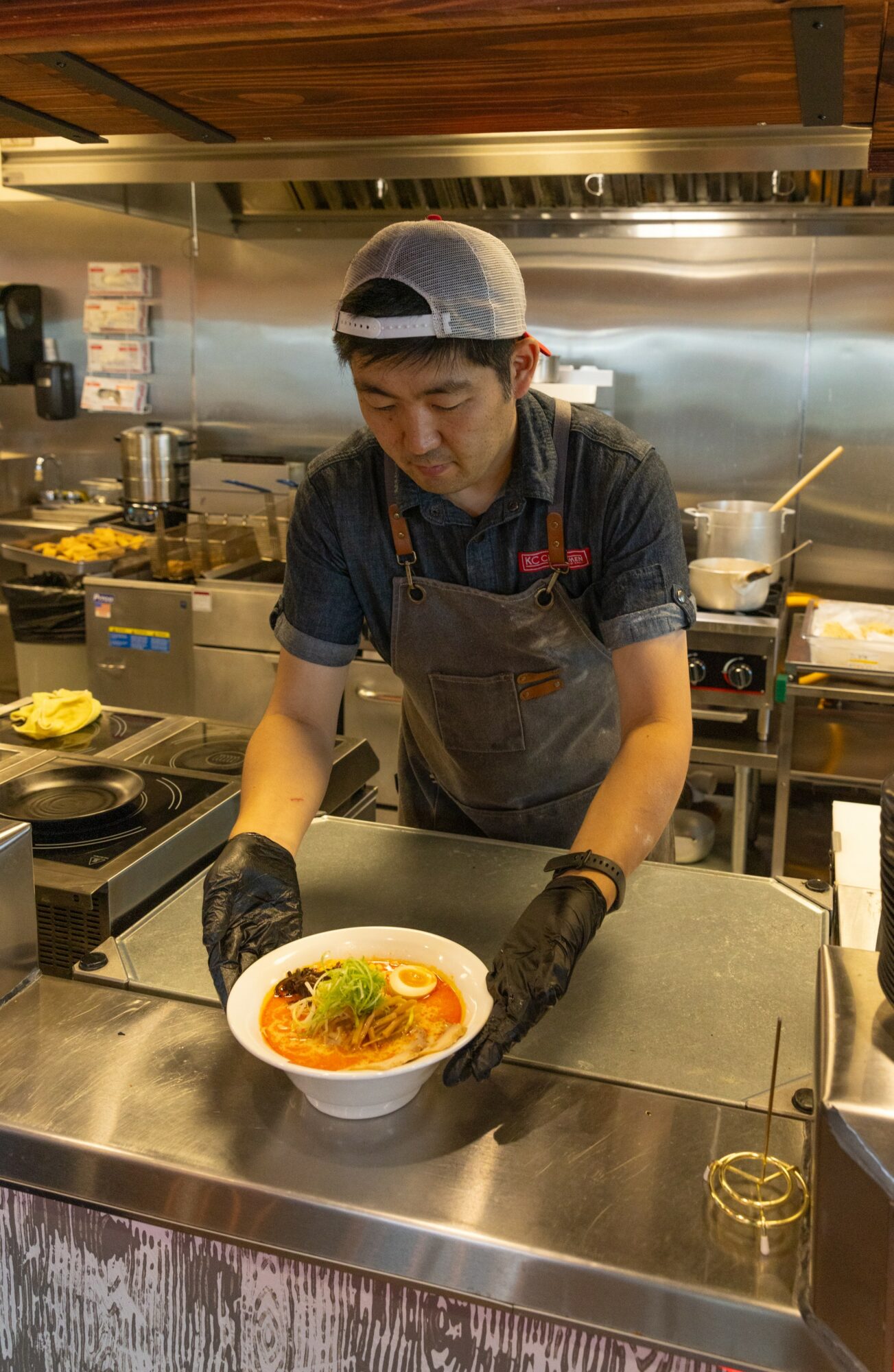 Person in a kitchen holding a bowl of ramen, wearing a cap, apron, and gloves, with cooking equipment in background.