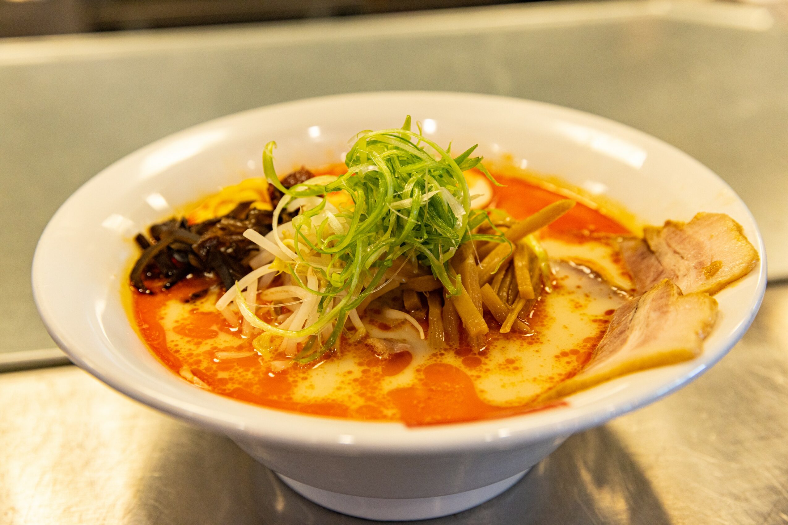 Bowl of ramen with broth, sliced pork, green onions, and vegetables, on a metal surface.