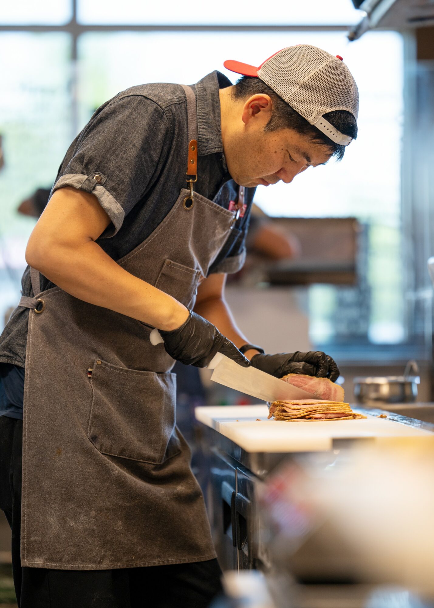 Young man wearing a cap, apron, and gloves preparing food in a kitchen, focused on his task.