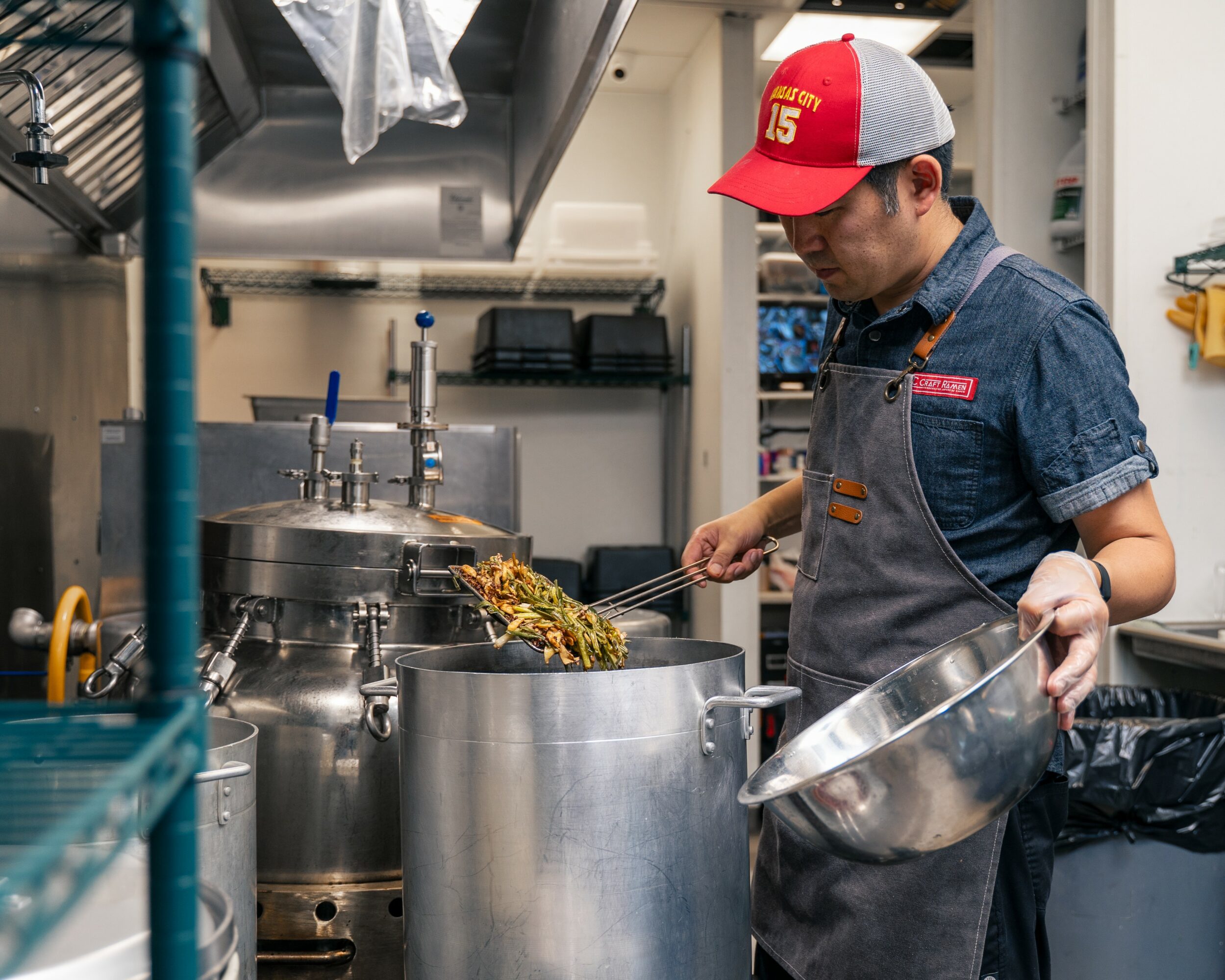 Person cooking in a commercial kitchen, stirring food in a large pot, wearing a red and gray cap and apron.