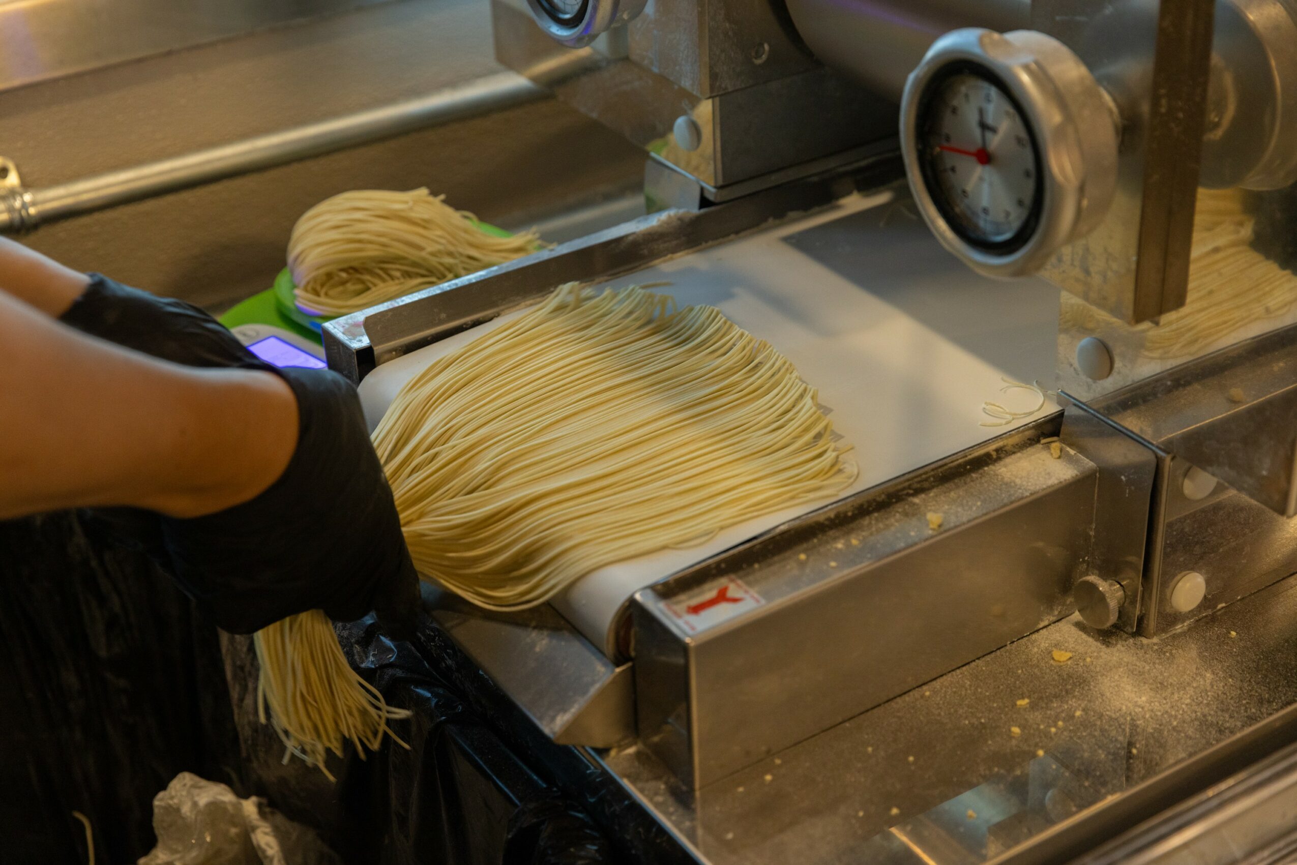 Person wearing black gloves handling freshly made pasta on a metal conveyor belt in a kitchen setting.