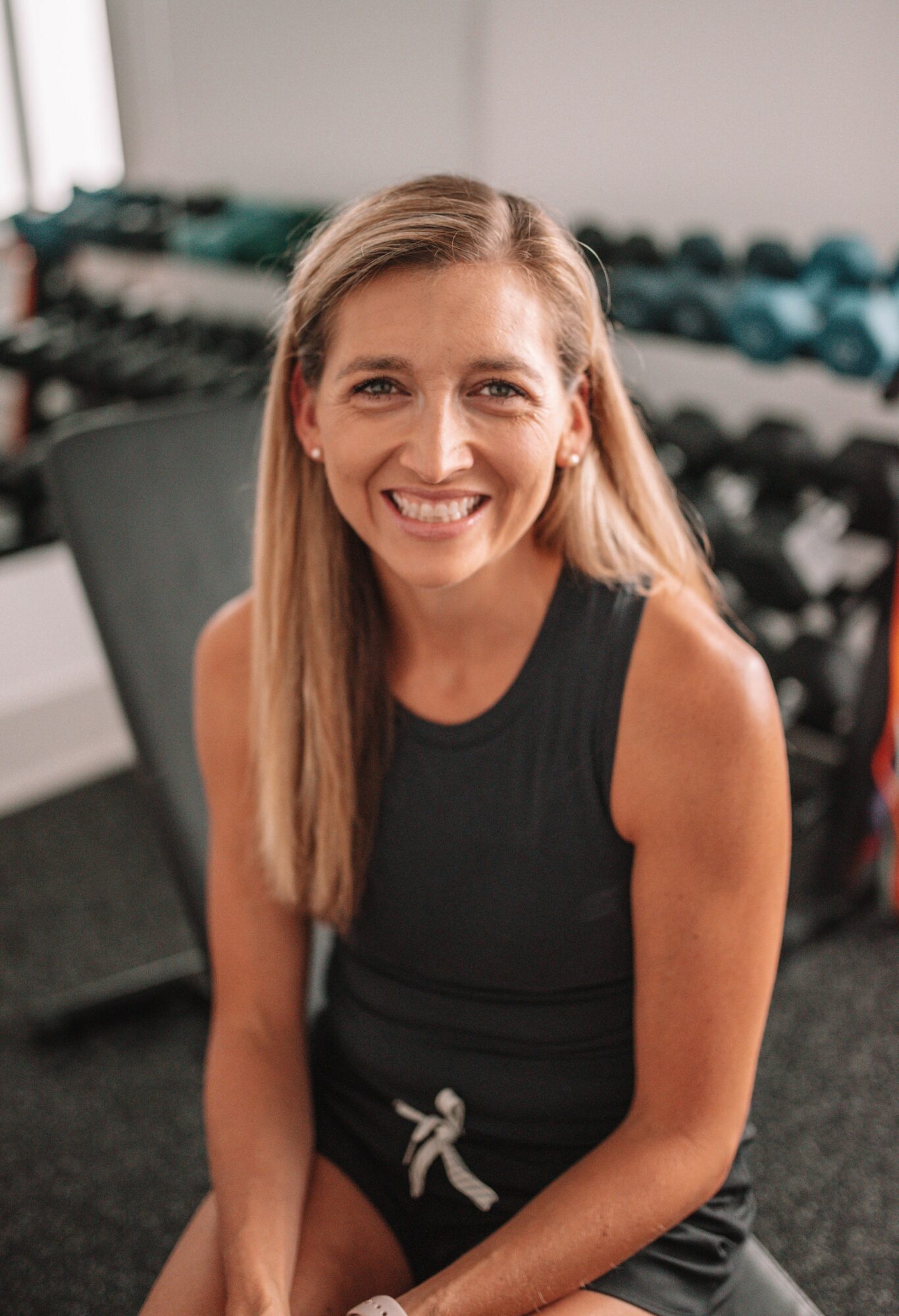 Smiling woman with long hair in a sleeveless black top, sitting in a gym with weights in background.