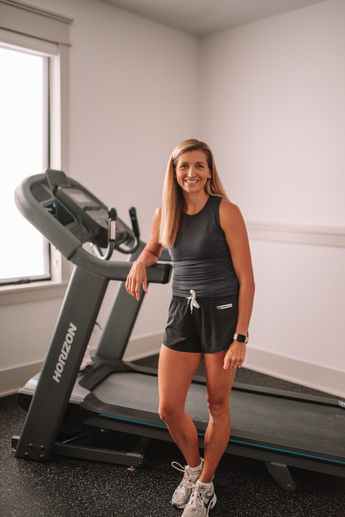 Smiling woman in workout clothes standing next to treadmill in gym room with window, white walls, and black flooring.