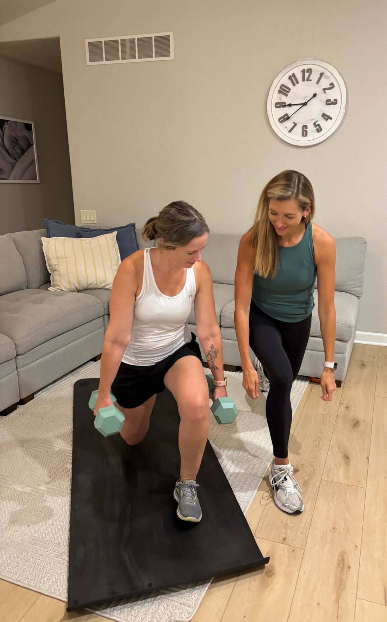Two women exercising on a black mat in a living room, one kneeling with dumbbells, the other standing nearby.
