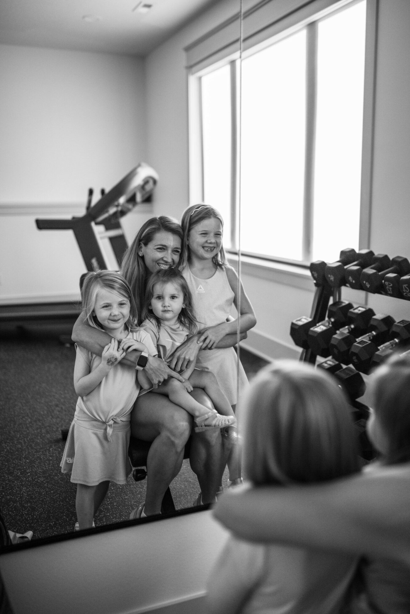 A woman with four young girls in a gym, some holding books, near a window and dumbbells, smiling.