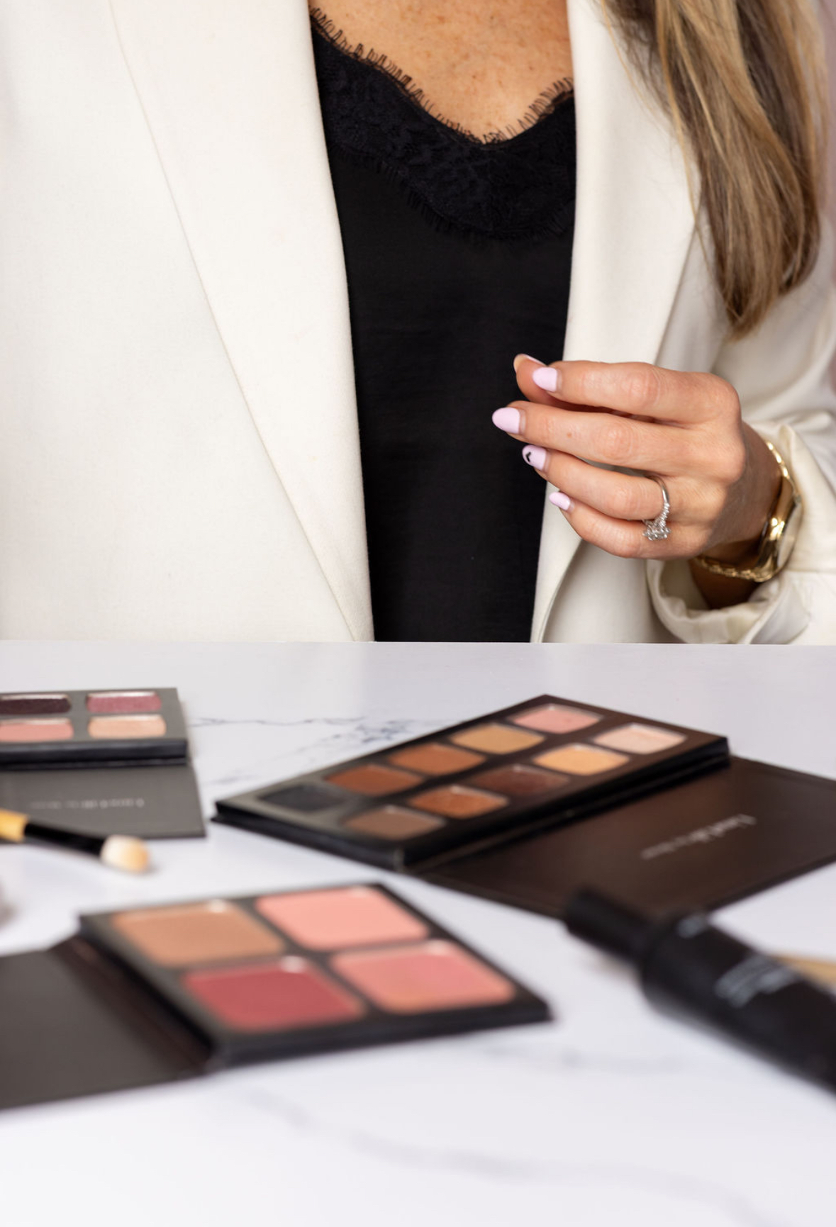 Close-up of a woman in a white blazer and black top, with makeup palettes on a table.