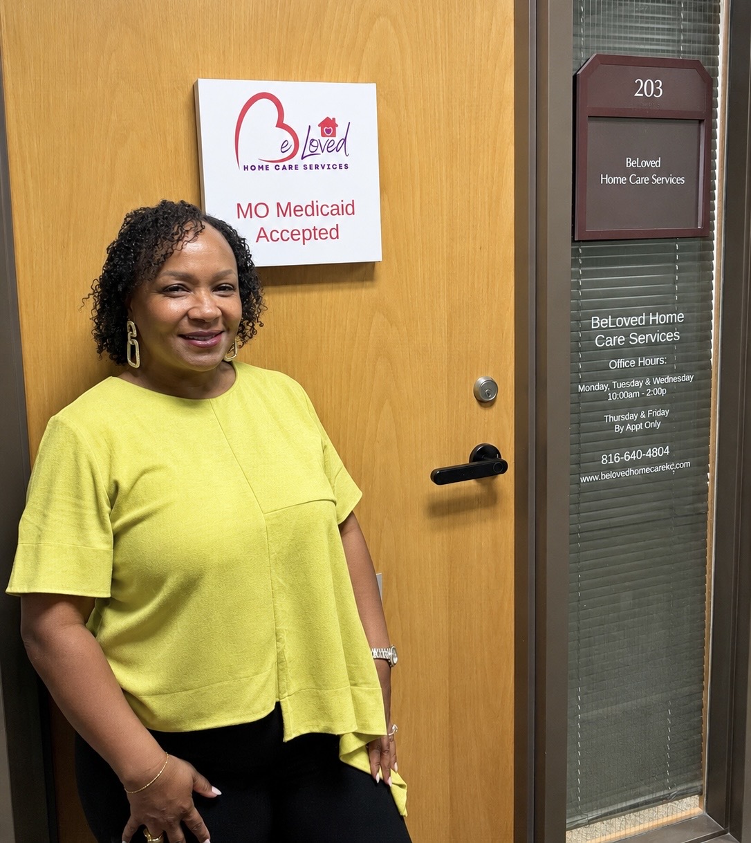 Woman with curly hair in yellow top standing next to door with signs, smiling, in an indoor setting.