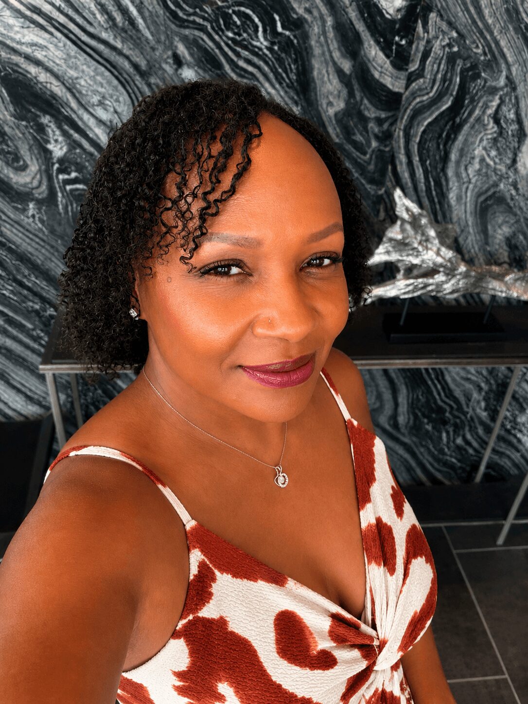 Woman with curly hair and makeup, wearing a patterned dress, taking a selfie indoors with a marble wall background.