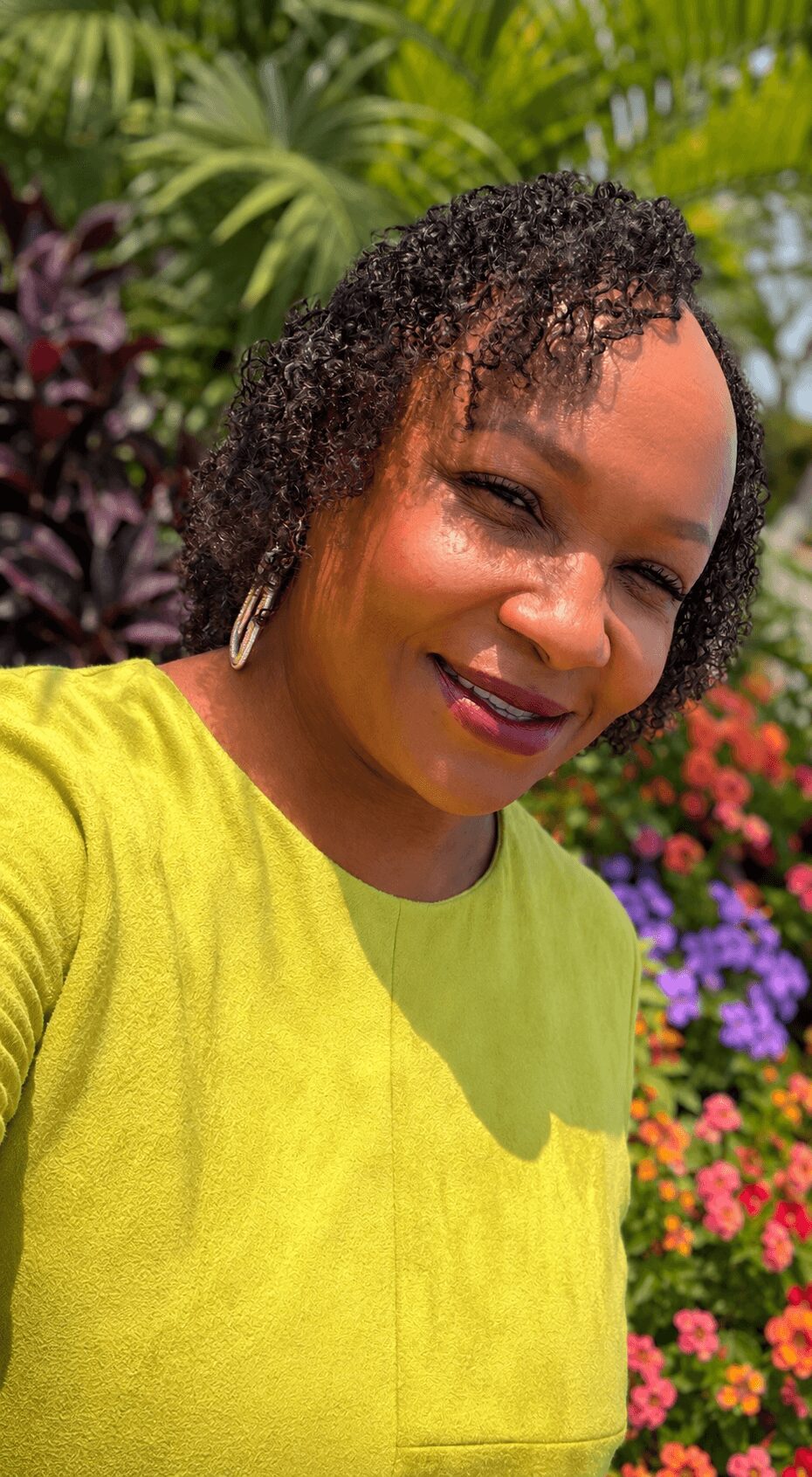 Woman with short curly hair smiling outdoors, wearing a yellow top, surrounded by colorful flowers and greenery.