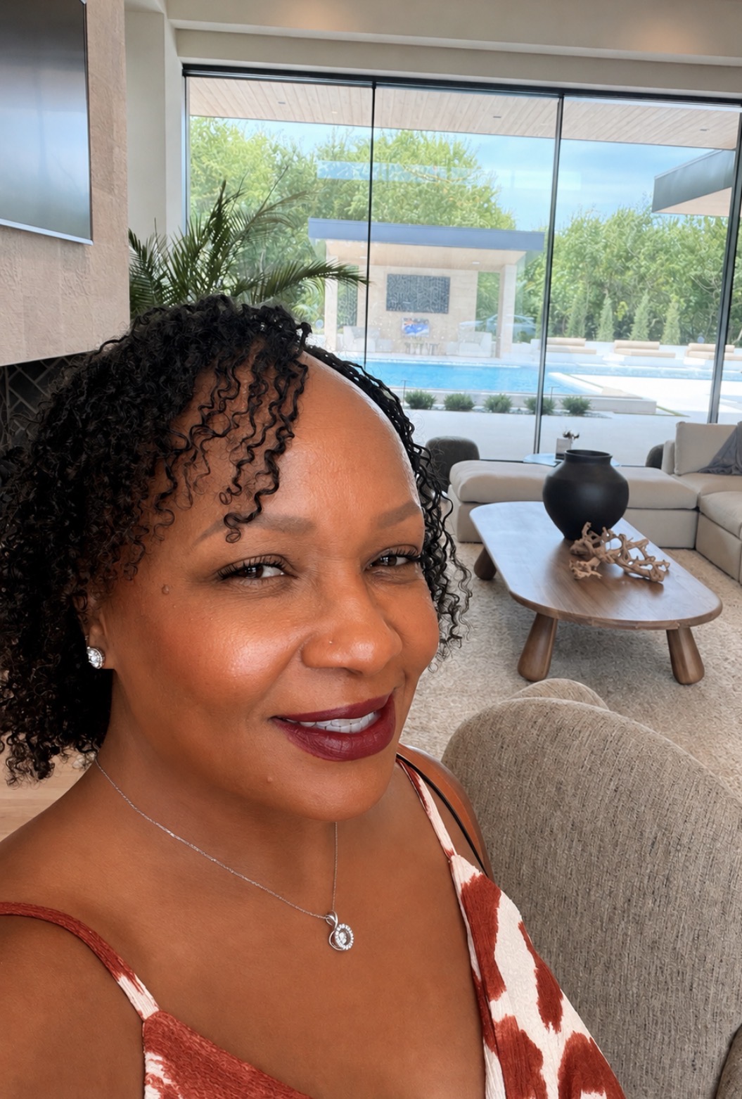 Woman with curly hair and dark lipstick smiling indoors, large window with trees outside in background.