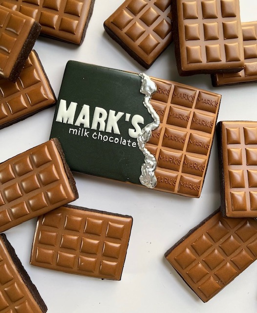 Unwrapped milk chocolate bar with pieces around it, and a partially unwrapped chocolate bar with a green wrapper labeled 'MARK'S milk chocolate'.