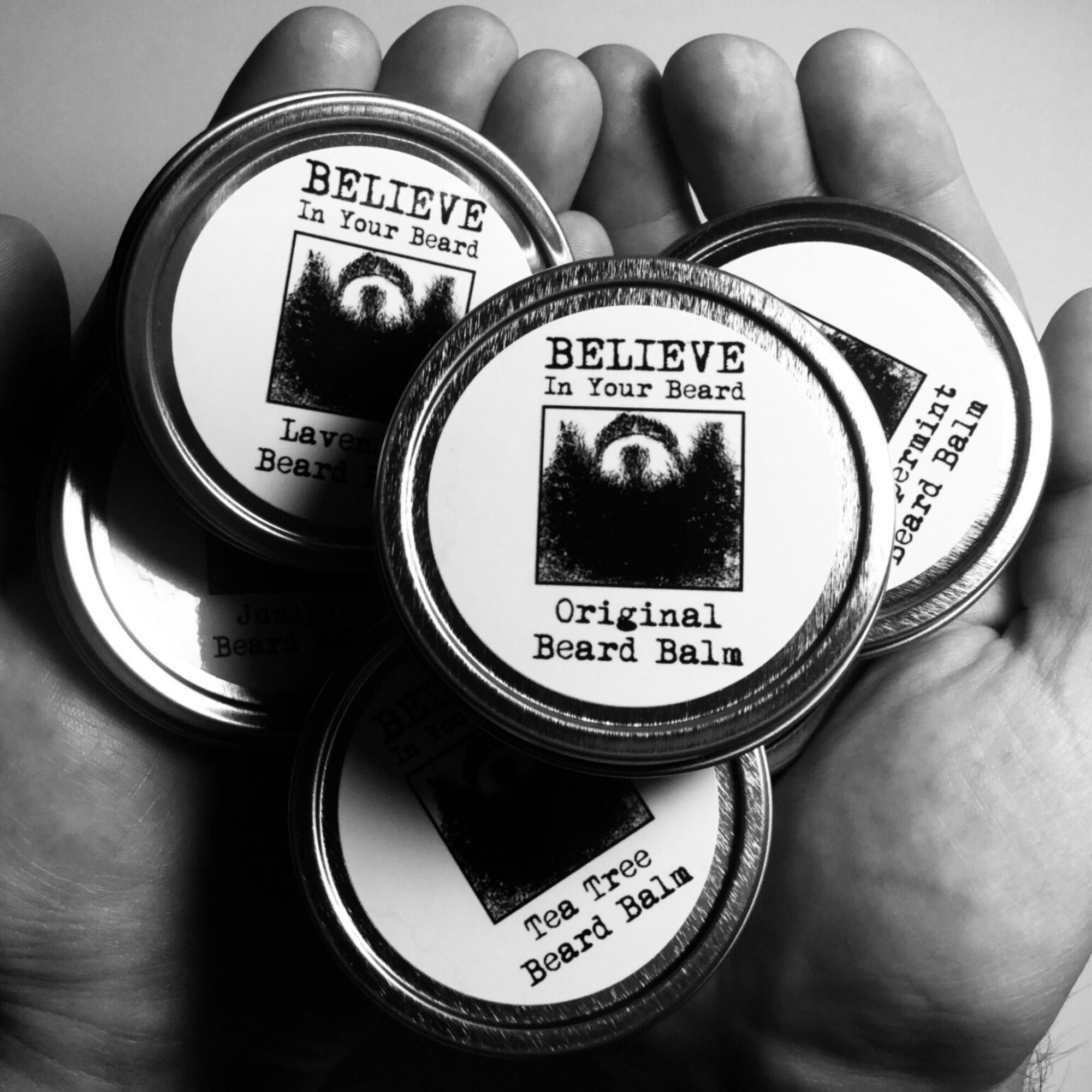 Hand holding three containers of beard balm with labels, in black and white.