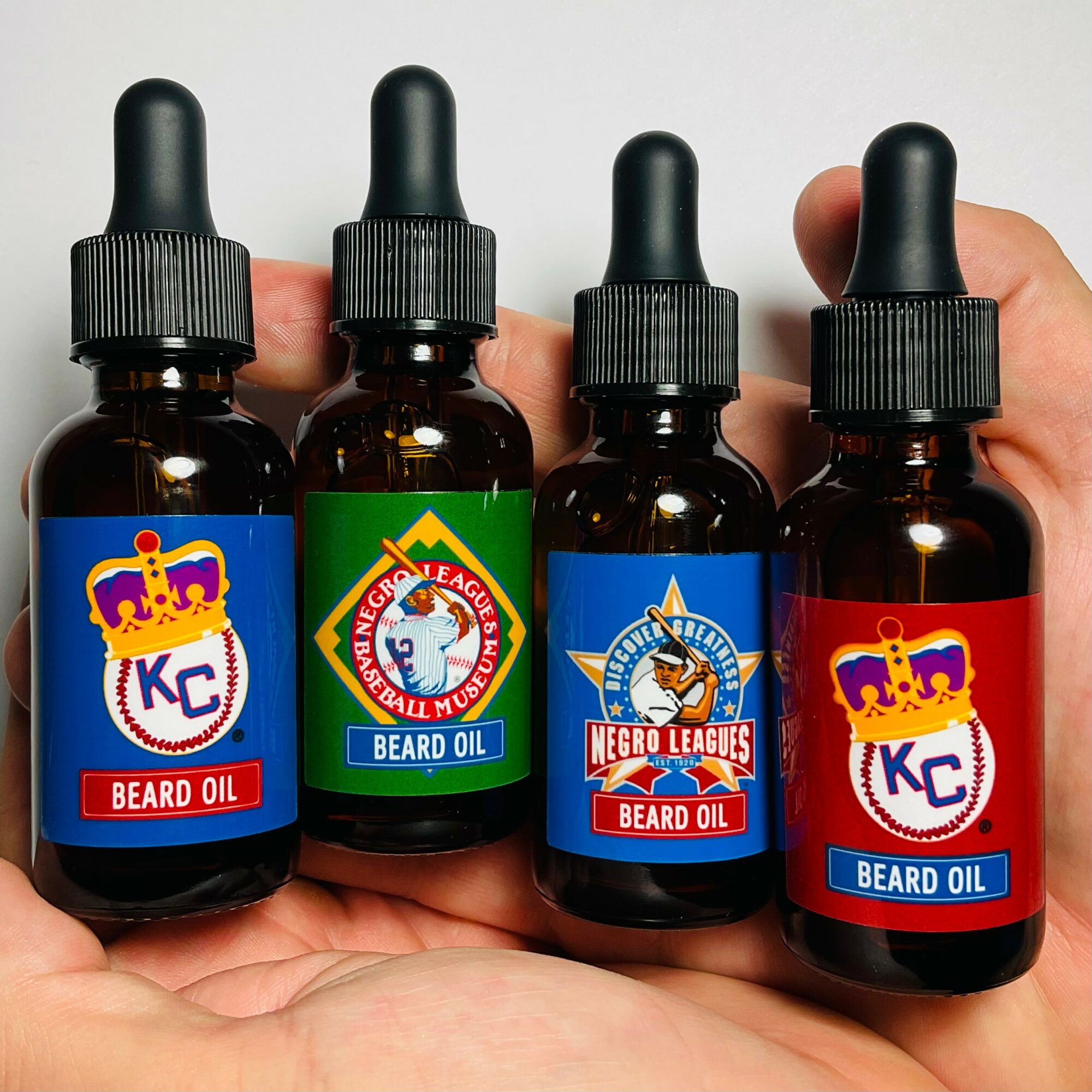 Four small bottles of beard oil held in a person's hand, each with a different label and logo.