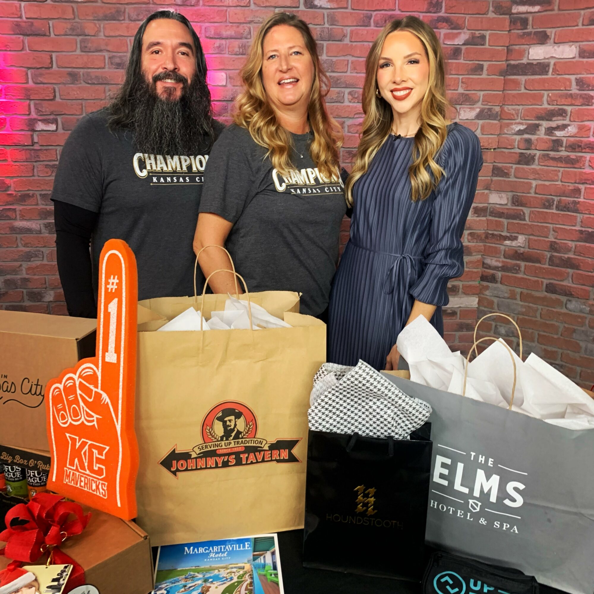Three people standing behind a table with shopping bags and a sign, smiling in front of a brick wall.