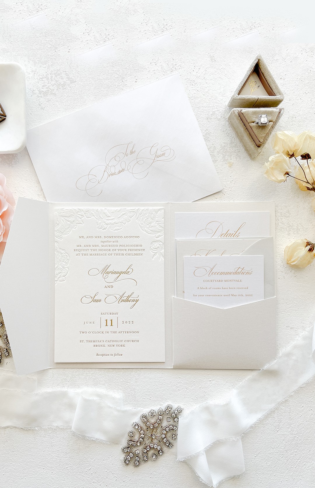 Wedding invitation with elegant script, surrounded by flowers, jewelry, and decorative items on a white surface.