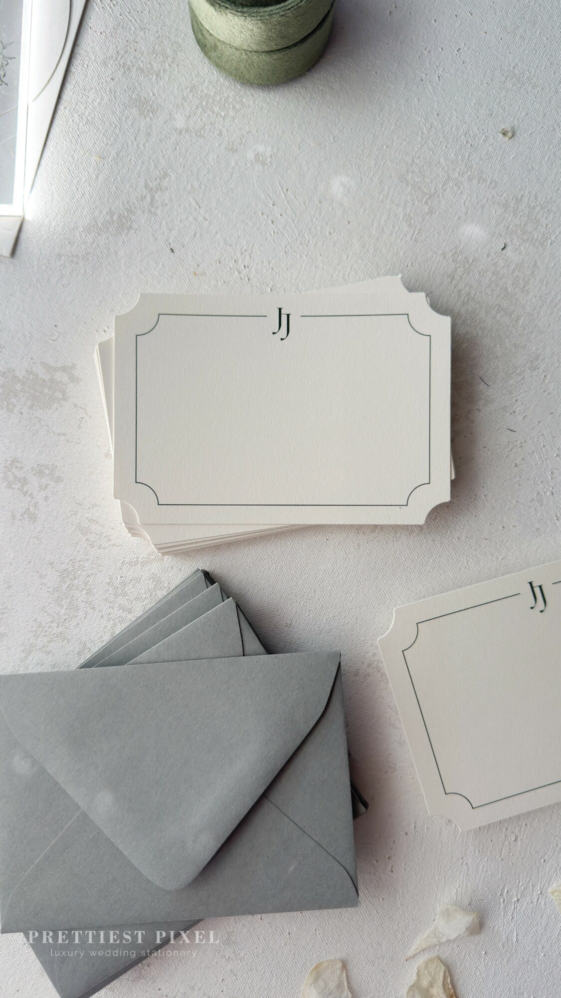 White card with decorative border, gray envelope, and another card on a light surface, with a green object nearby.